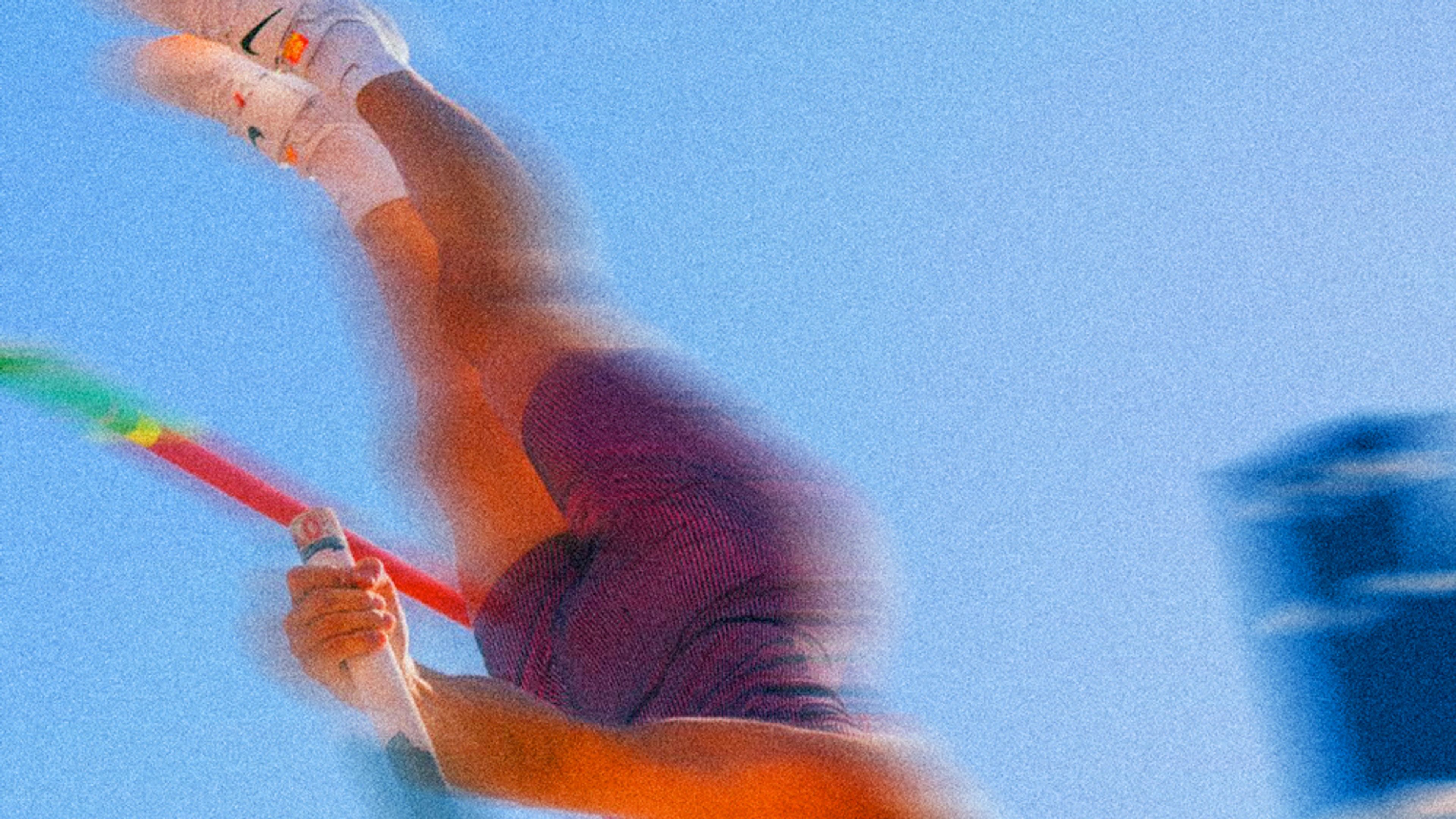 A blurred image of a pole vaulters legs as they clear the bar. You see the top of the pole, and white Nike shoes in the air, against a blue sky.