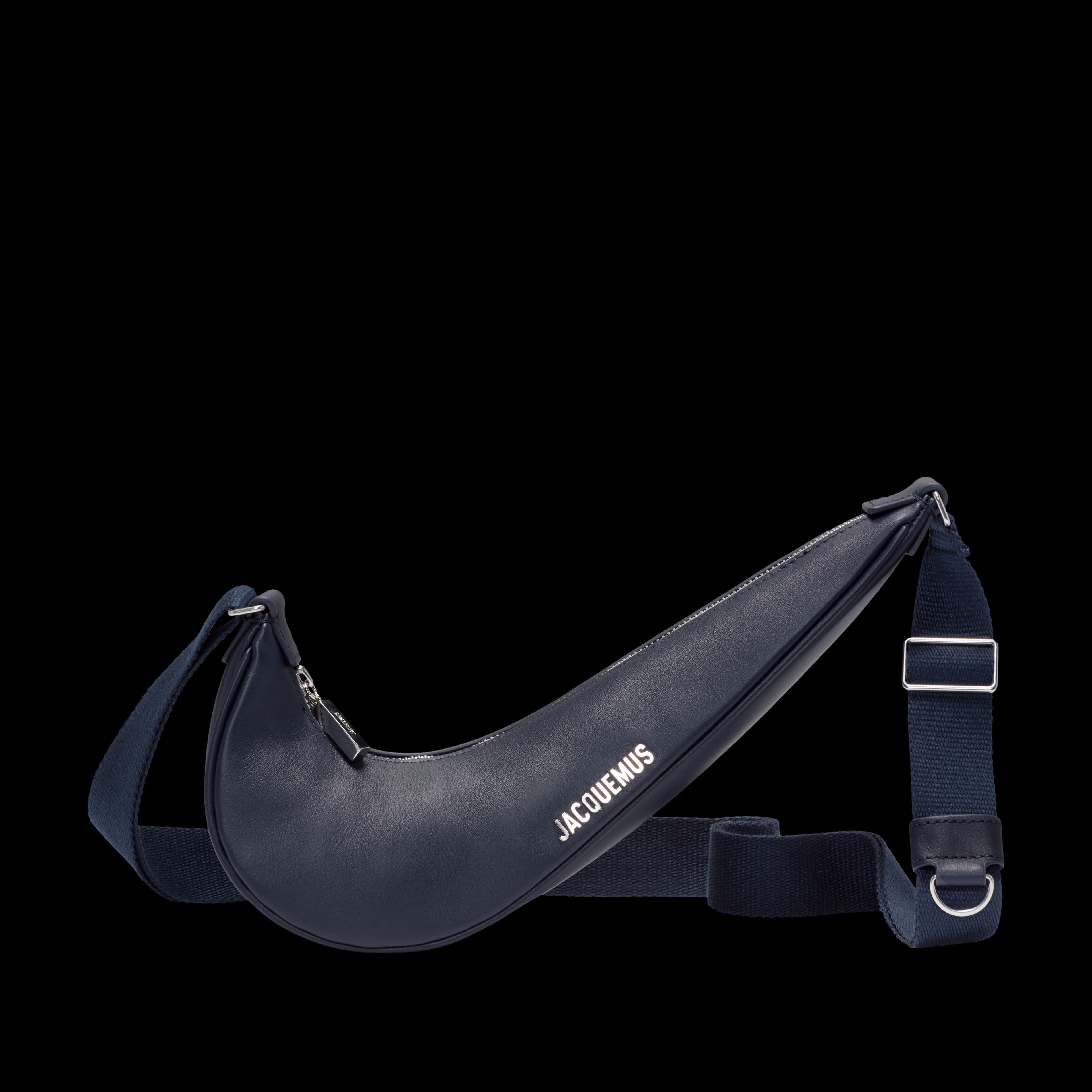 Front view of the Nike x Jacquemus Le Swoosh Crossbody Bag in midnight navy, with bright silver bag hardware and Jacquemus brand mark. The bag itself is in the shape of a Nike Swoosh, and includes a navy blue strap that is draped behind the body of the bag. The bag floats on a black background.