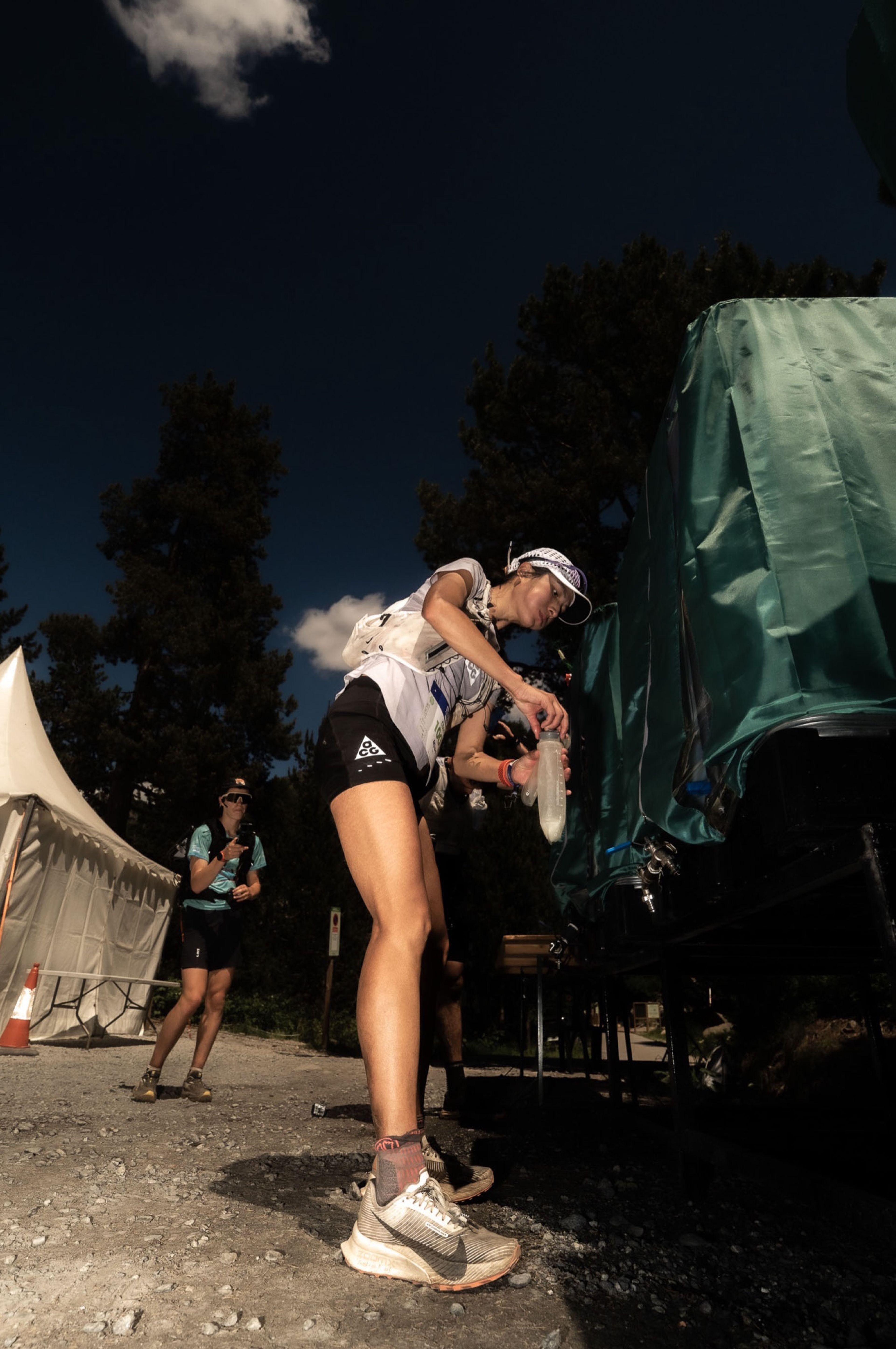 Yao Miao, Chinese trail runner, refills water bottles at an aid station during an ultra-endurance race, wearing Nike ACG trail gear.