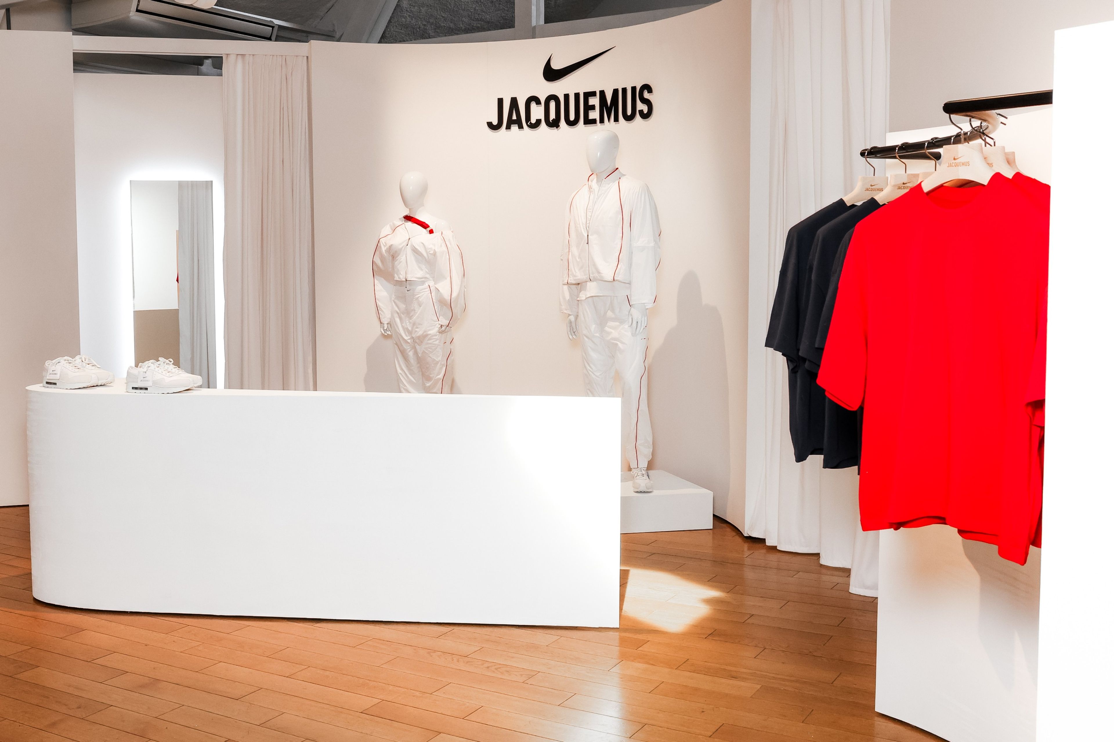 View into the Jacquemus Station at the Nike Athlete House in Paris, France. We see the Nike X Jaquemus men's and women's track suits on white mannequins. Nike x Jacquemus short sleeve tees in Univserity red and black hang on a bar to the right. A curved white counter holds the Nike x Jacquemus Air Max 1 Sneaker in white. Fitting rooms can be seen in the background.