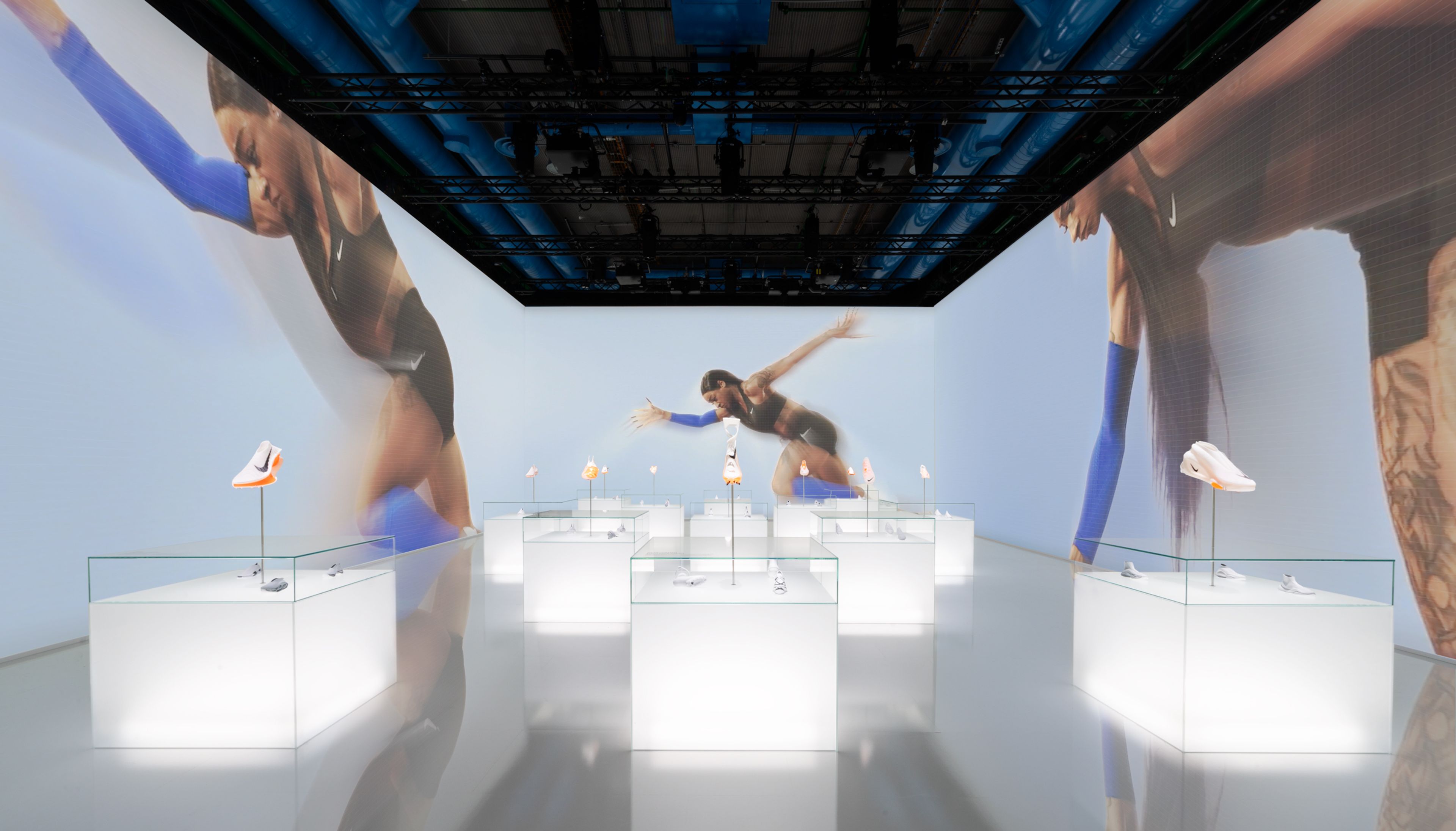 Inside of Gallery 4, within the Nike Au Centre Pompidou, there are a dozen glowing white display cabinets showcasing innovative Nike footwear featuring Air technology. Images of Sha'Carri Richardson in different phases of a sprint, appear on 3 walls of the room.