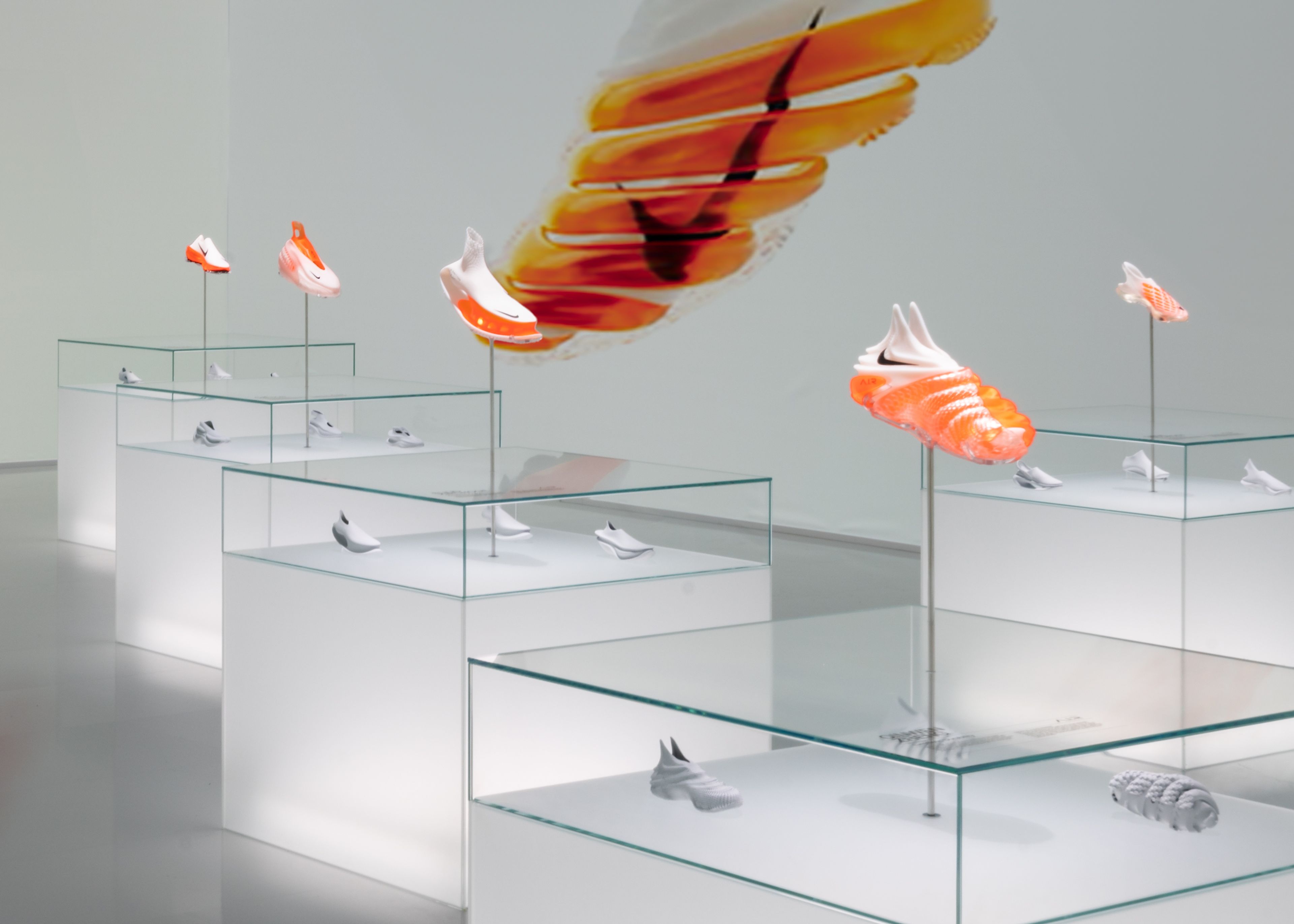 Detail view of the glowing display cabinets inside of Gallery 4, Nike Au Centre Pompidou. A single footwear design sits above each glass cabinet, with other footwear and Air technology elements displayed inside. All product is white with black Swoosh, featuring bright orange Air units. The wall behind the cabinets showcases a large image of an innovative design with a bright orange Air unit spiraled completely around the shoe.