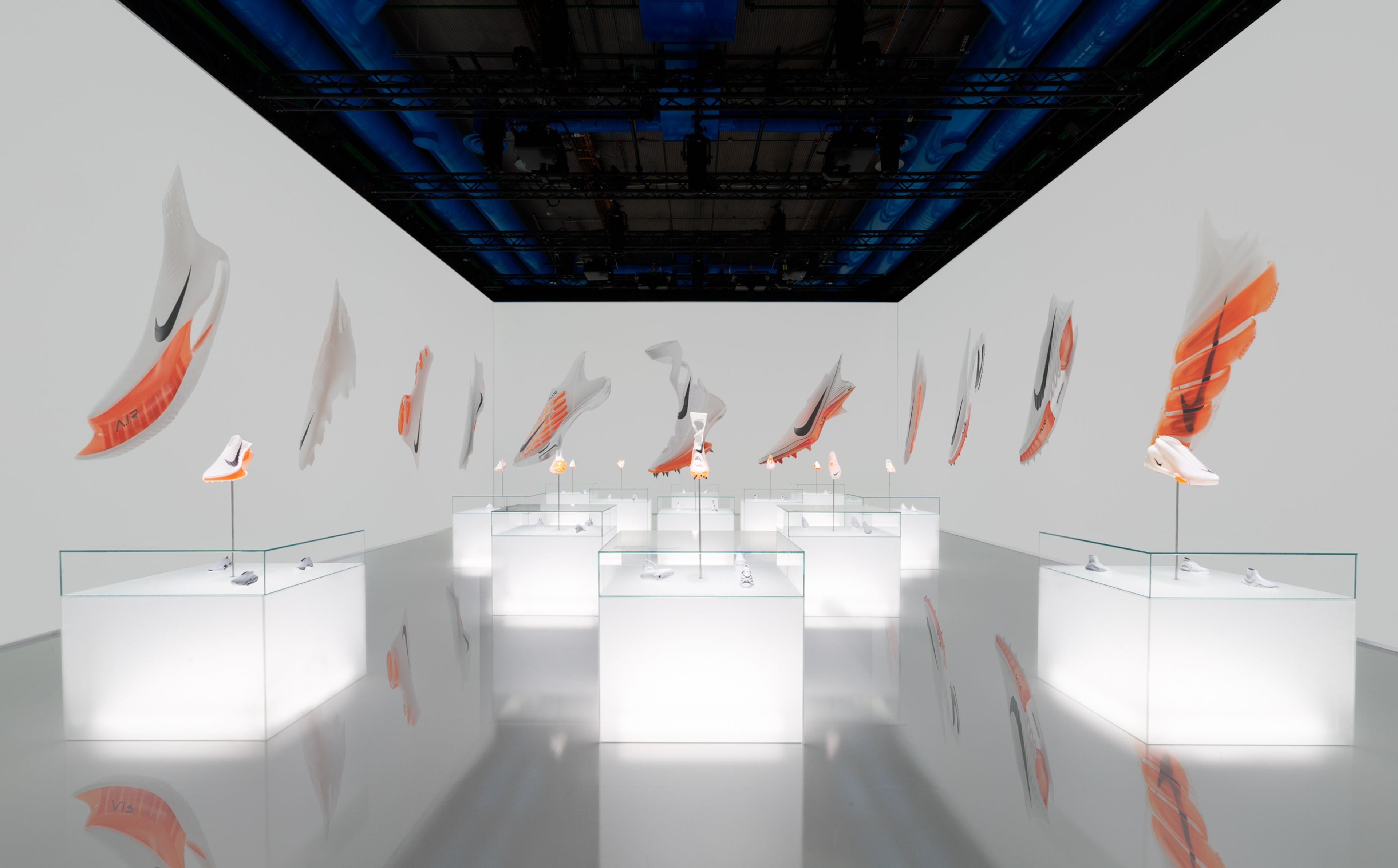 Inside of Gallery 4, within the Nike Au Centre Pompidou, there are a dozen glowing white display cabinets showcasing Innovative footwear designs featuring Air technology. All products are white with a black Nike Swoosh, and Air units are in bright orange. On the 3 walls, we see blown up photos of innovative designs floating against a white background.