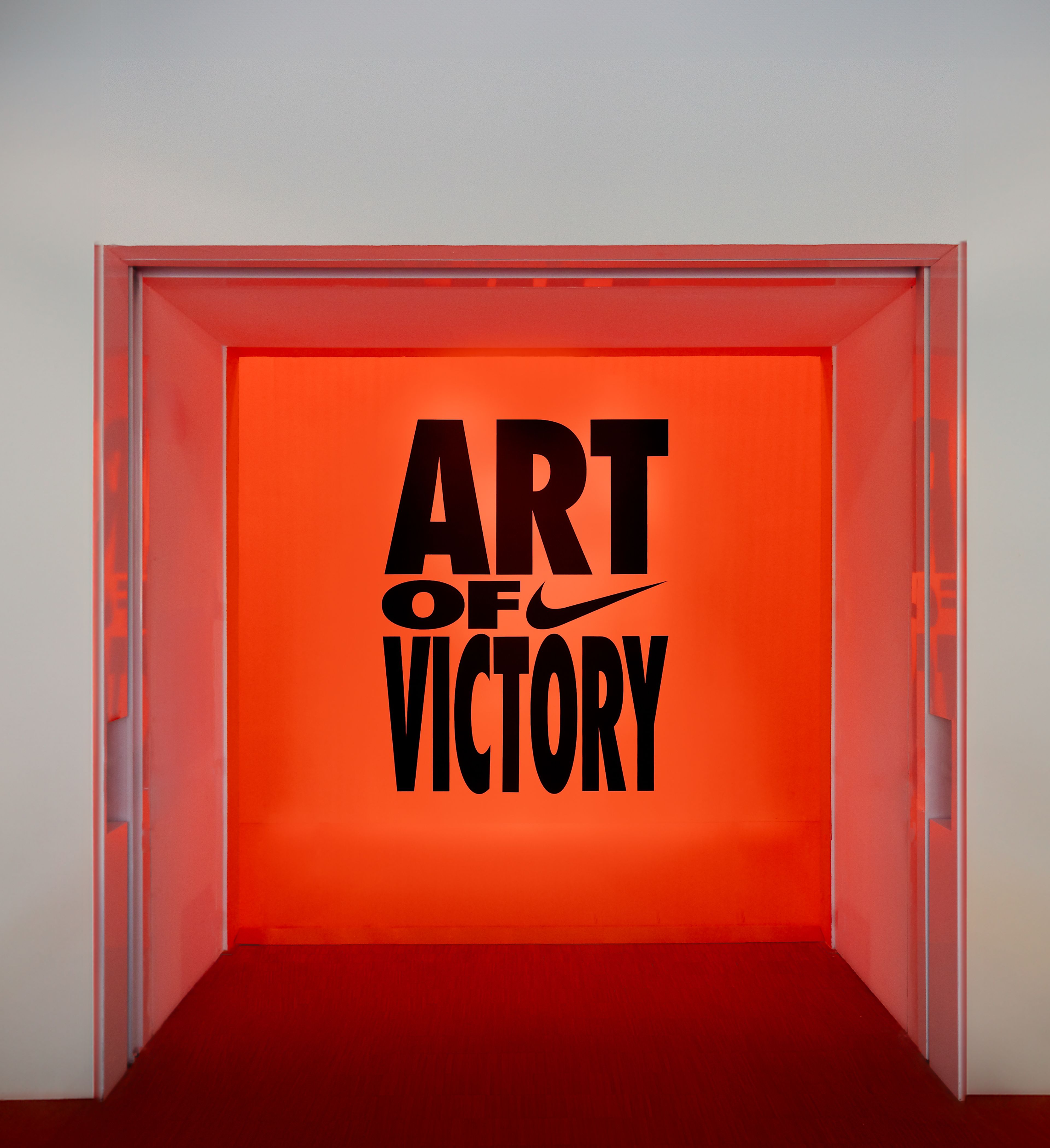 Detail view of a wall within the Art of Victory exhibit in Gallery 4 of the Nike Au Centre Pompidou, located in Paris, France. The glowing orange wall showcases the words Art of Victory in bold, black lettering along with a black Nike Swoosh.