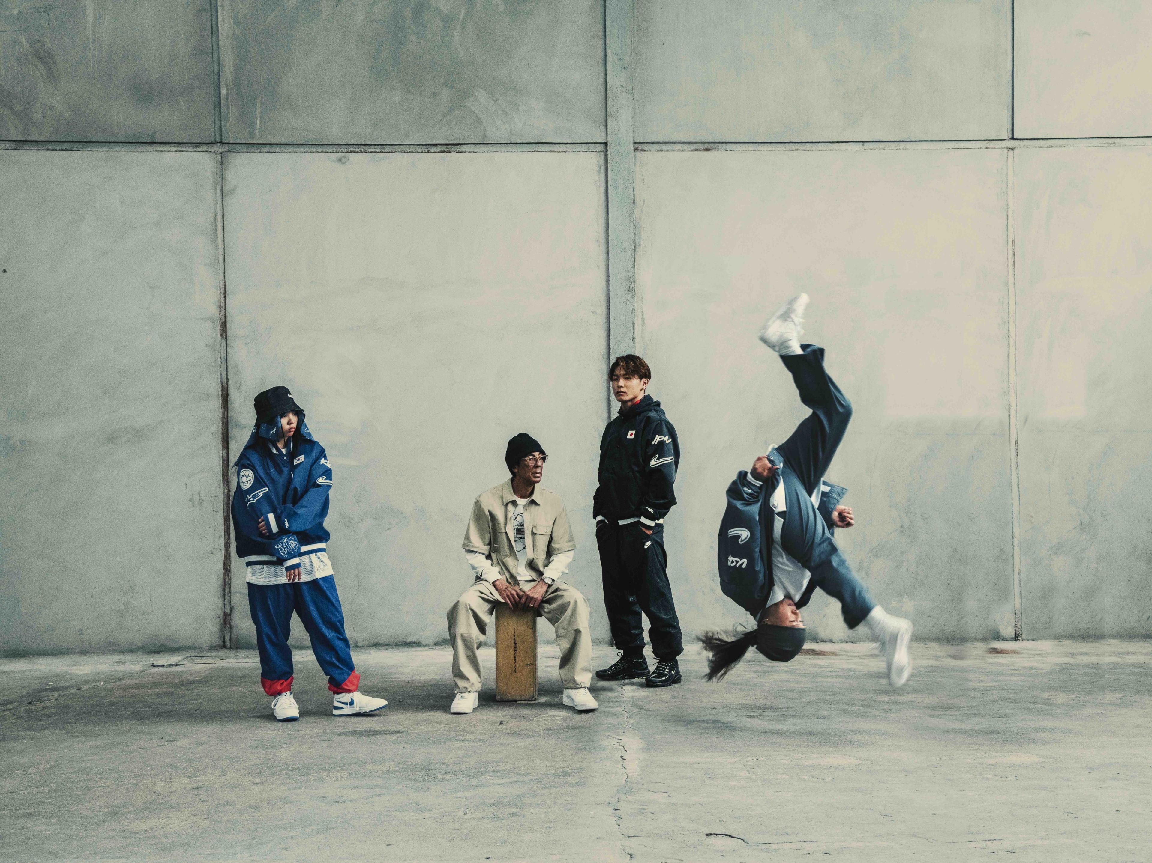 From left to right, JiYun Shun, Futura, and Shigeyuki Nakari watch as Sunny Choi does a front flip during a breakdance in white Nike Jam shoes. The breakdancers are located in a concrete space with grey floors and walls, and all wear attire from the Nike Breaking Collection.