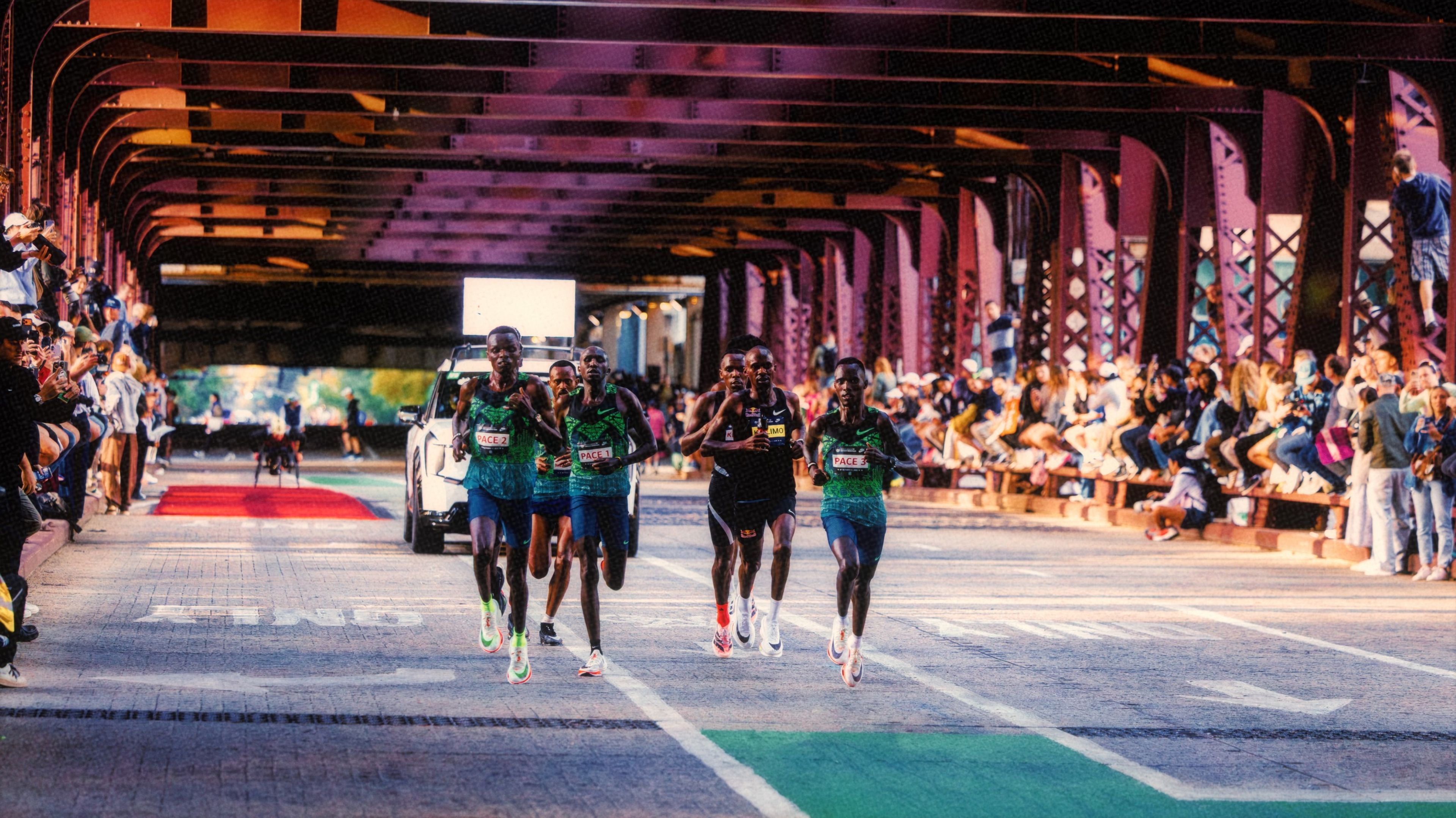 Elite male runners, including Jacob Kiplimo, wearing Nike racing kits compete on a Chicago street beneath a steel bridge with spectators cheering from both sides.