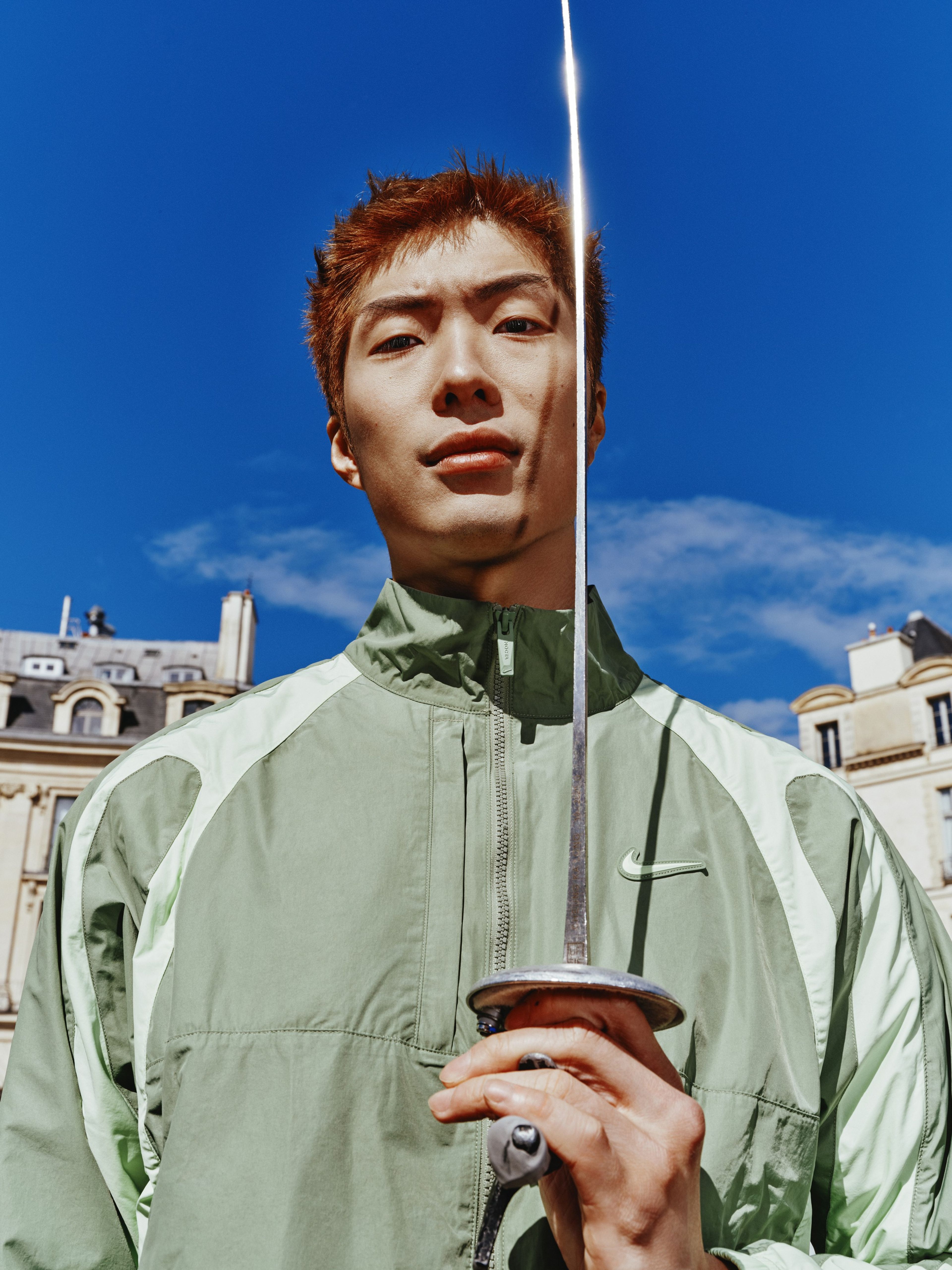 Cheung Ka Long holds his fencing foil upwards in front of his face, while looking directly at the camera. We see only the top half of his body, and he is wearing the NOCTA Northstar Nylon Track Jacket in Oil Green. There is a bright blue sky behind him, and glimpses of a couple of buildings.