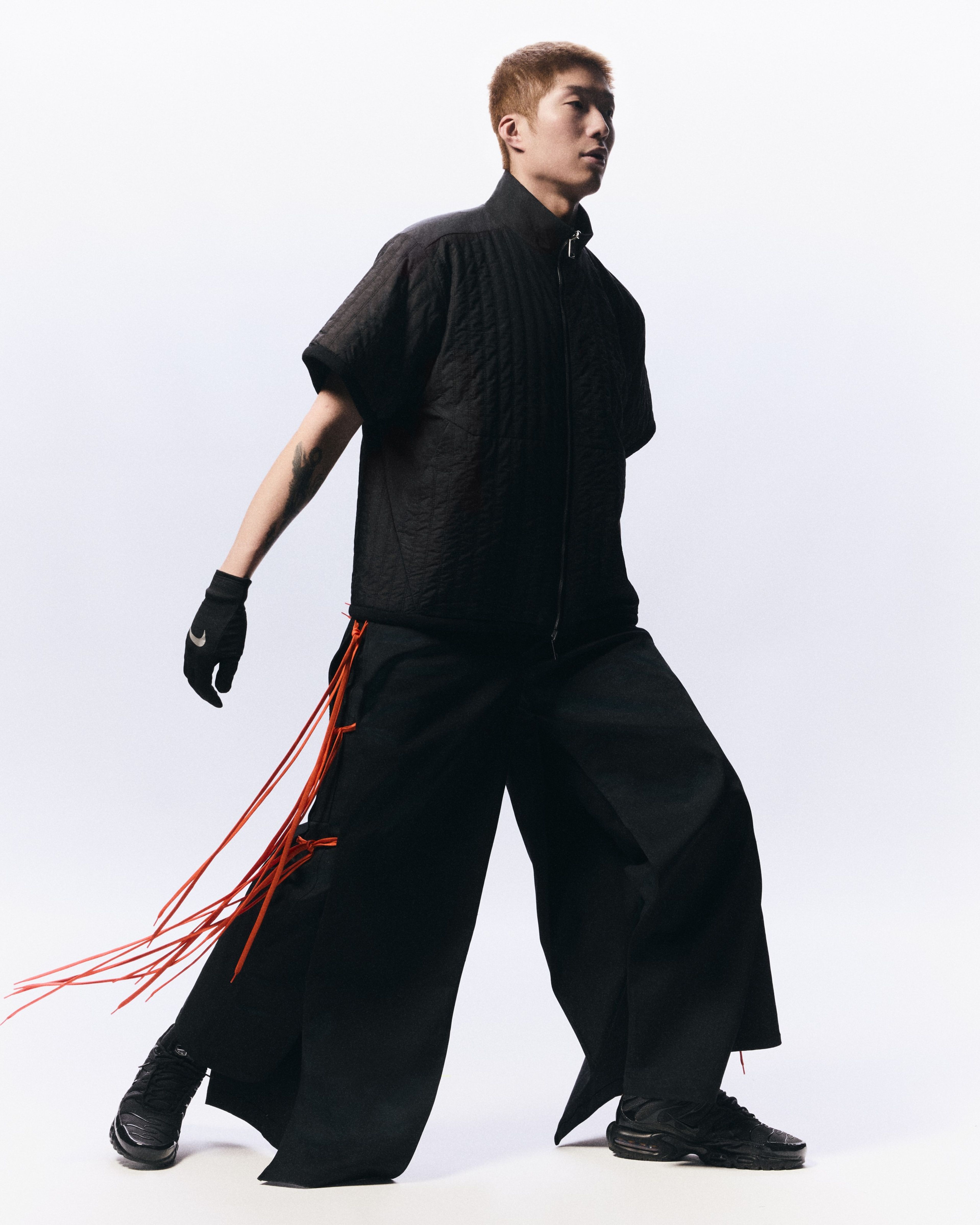 Full body portrait of Cheung Ka Long as he strides across a white background. He wears oversized black pants with red ties, a black short-sleeve zip up shirt, black Nike footwear, and black gloves with a reflective Swoosh on the back of the hand.