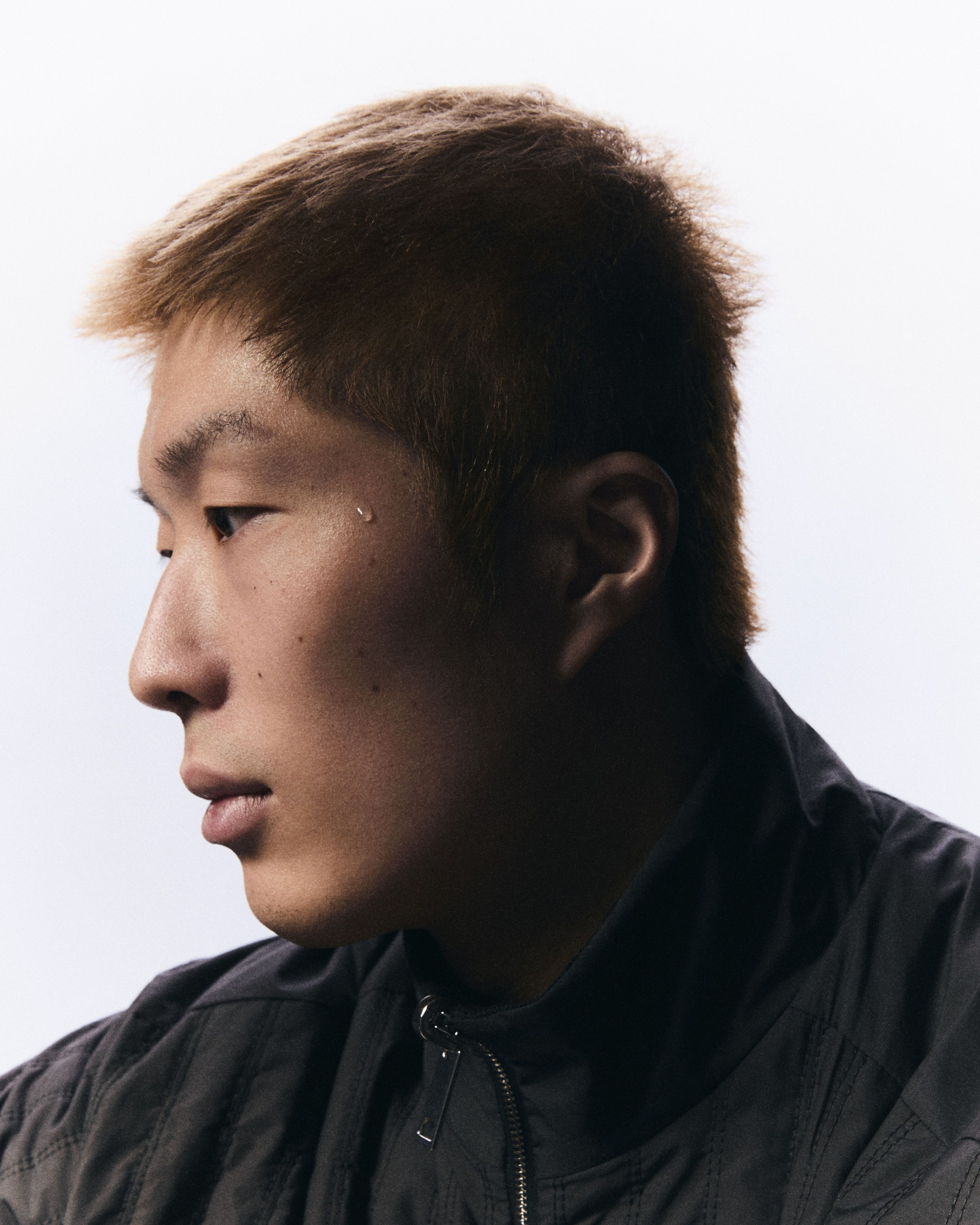 Side view portrait of Cheung Ka Long, facing to the left. His face is framed against a white background, and he wears a black jacket. His face glows in the light, while his neck and ear is in shadow.