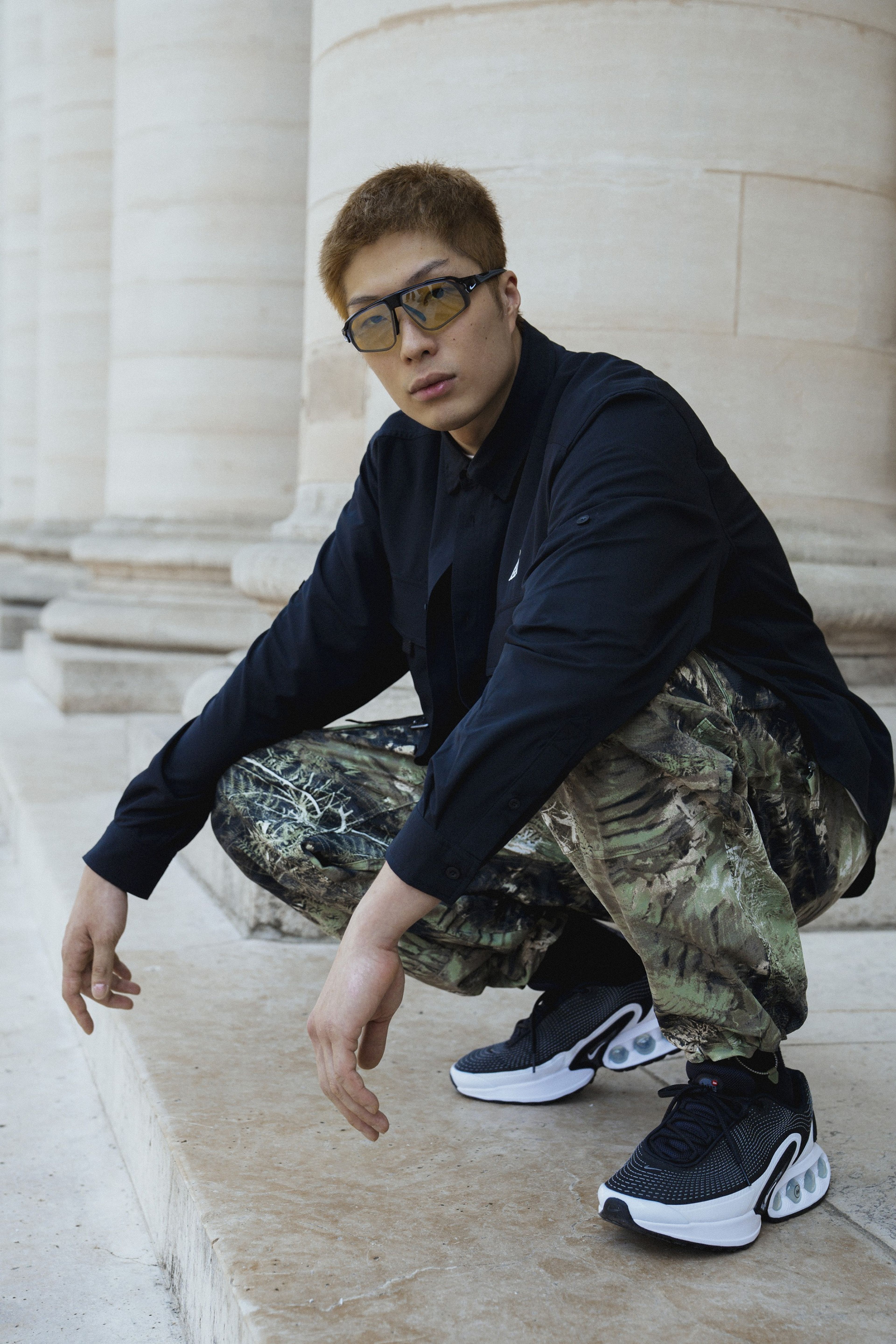 Full body portrait of Cheung Ka Long, crouching in front of large stone pillars. He is wearing a pair of Air Max DN shoes, Nike ACG "Smith Summit" pants with a green all-over camo print, and a black Nike ACG Dri-FIT UV "Devastation Trail" long sleeve shirt. Long looks directly at the camera, wearing a pair of Nike glasses with wrapping lenses and bold black frames.
