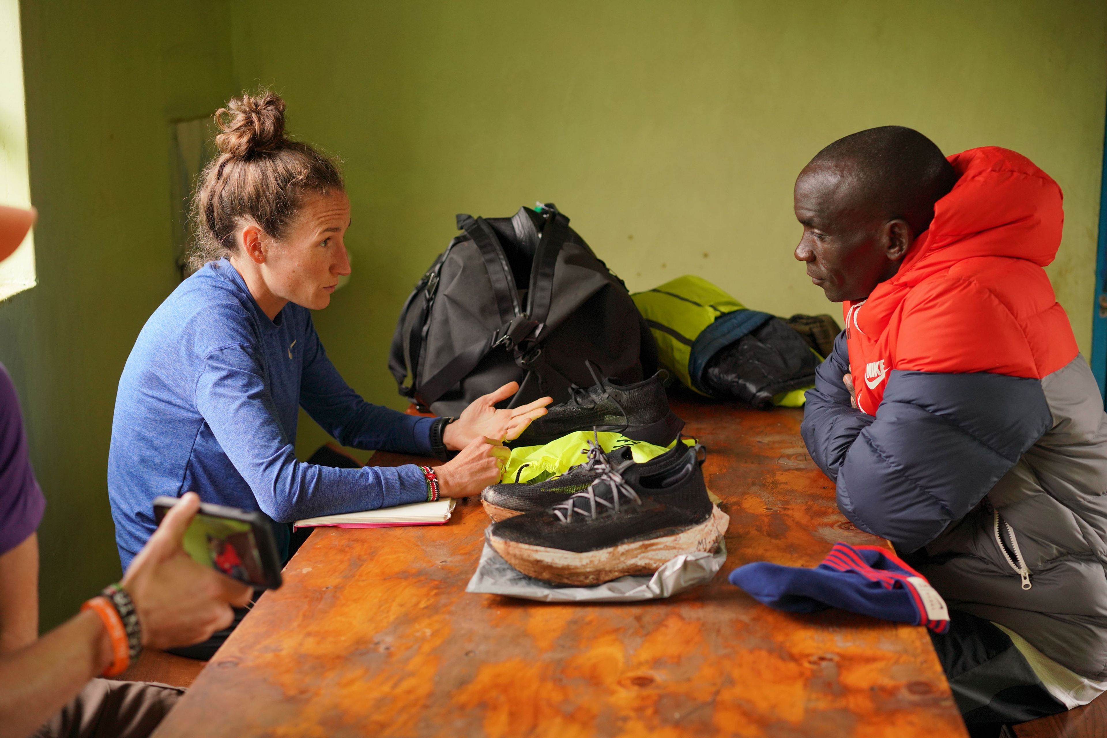 Carrie Dimoff and Eliud Kipchoge discuss Nike running shoes at a wooden table, with duffel bags and a notebook nearby, indoors.