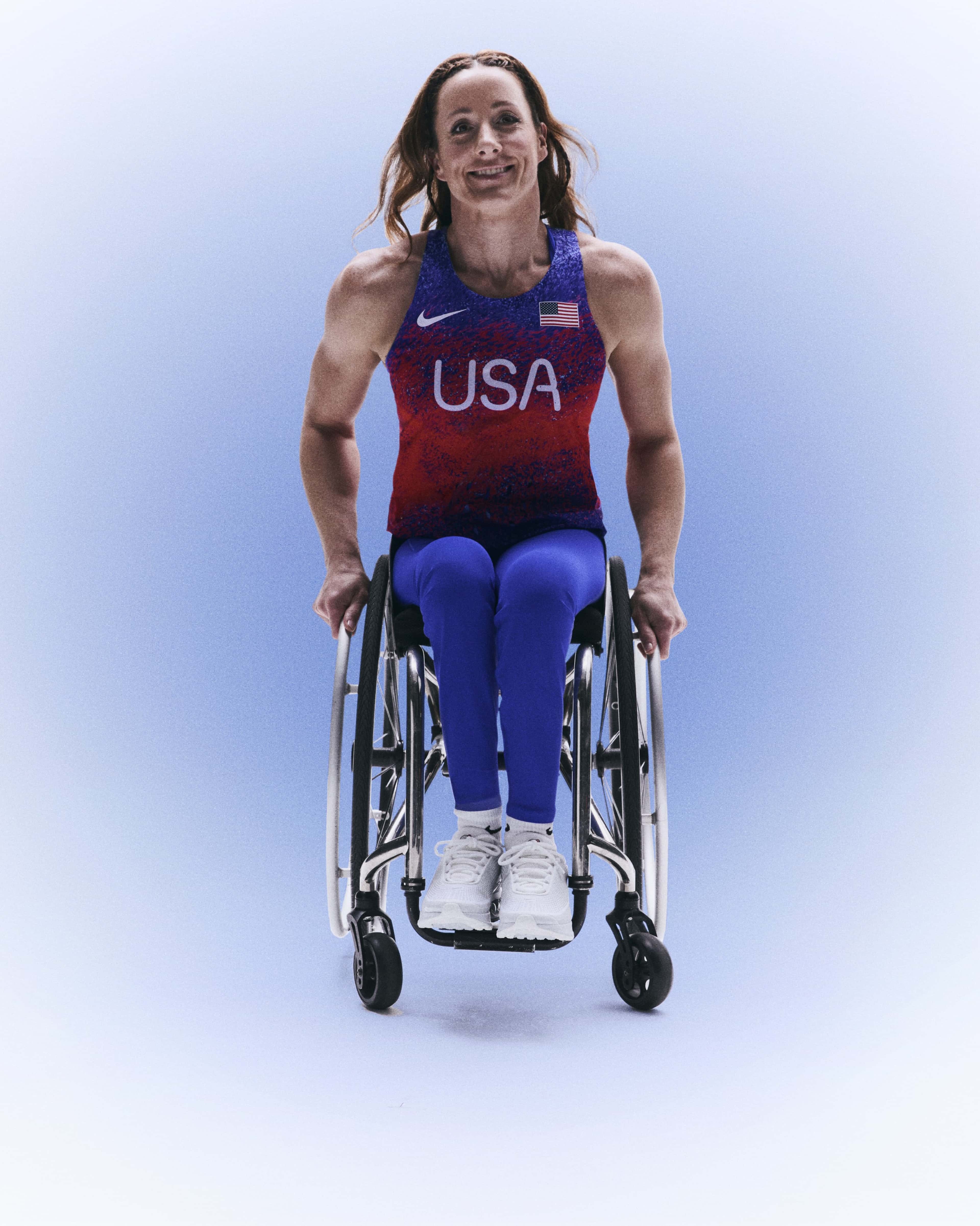 Full body portrait of Tatyana McFadden wearing her Team USA track and field uniform, with her hands resting on the hand rails of her wheelchair. Tatyana smiles directly at the camera with her hair blowing slightly back, against a blue gradient background.