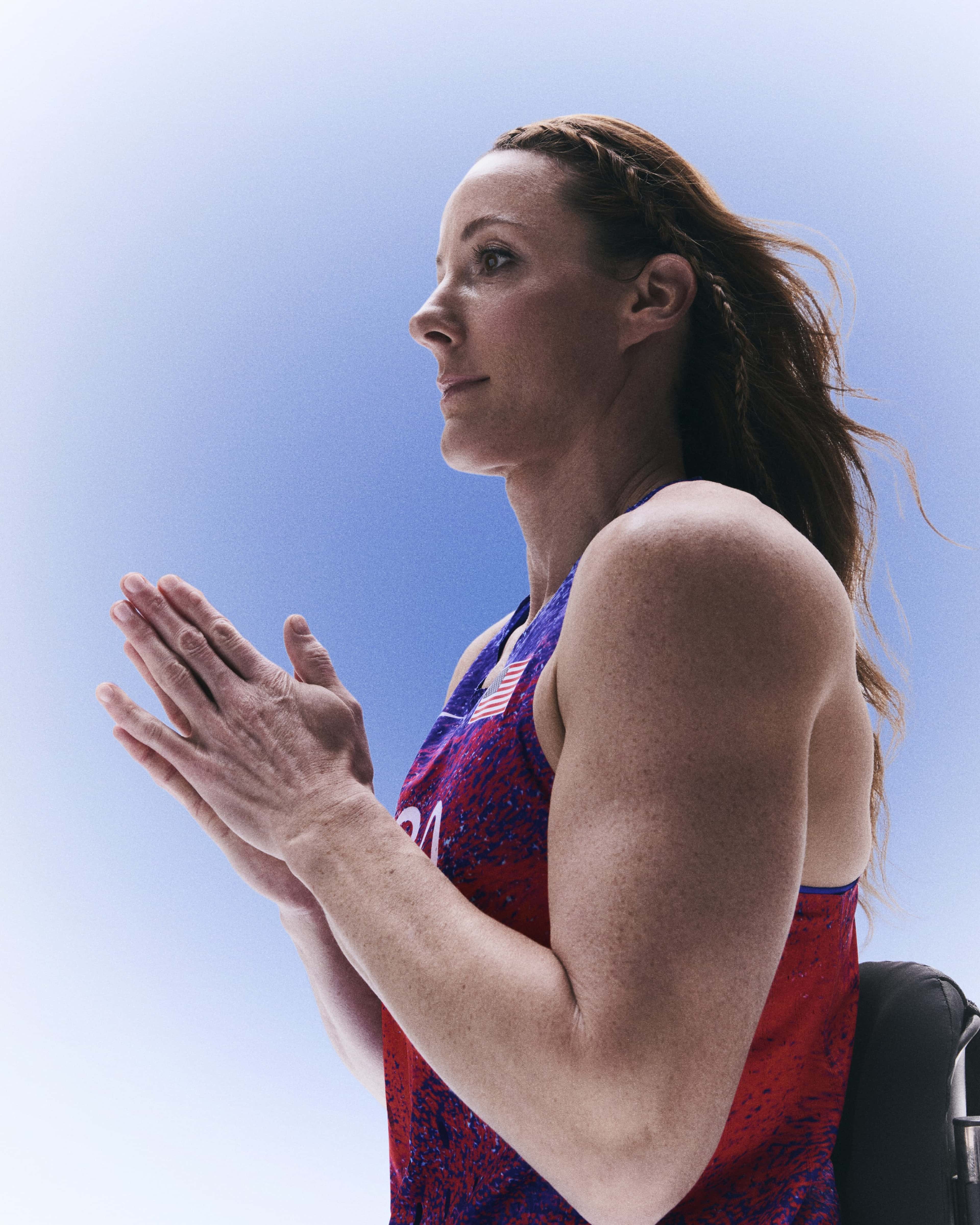 Side view of Tatyana McFadden pressing her hands together, while facing left. Tatyana wears the Team USA track and field singlet, and her hair is loose with a single braid across the front. She appears against a blue gradient background.