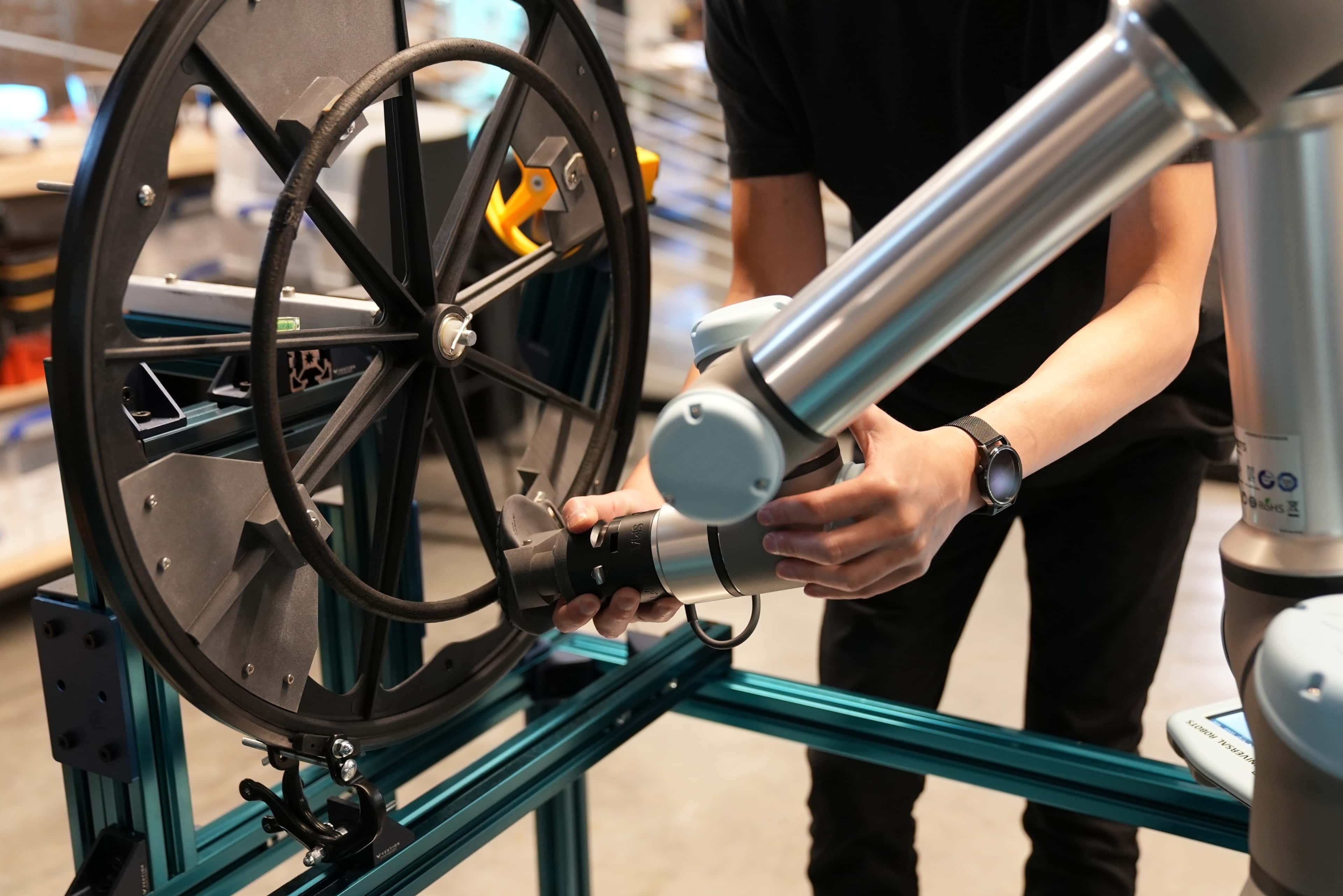 A member of the research team adjusts the robot mechanical arm during testing in the Nike Sports Research Lab, in Beaverton, OR. We see a close up of two hands adjusting the silver arm against the black wheelchair wheel.