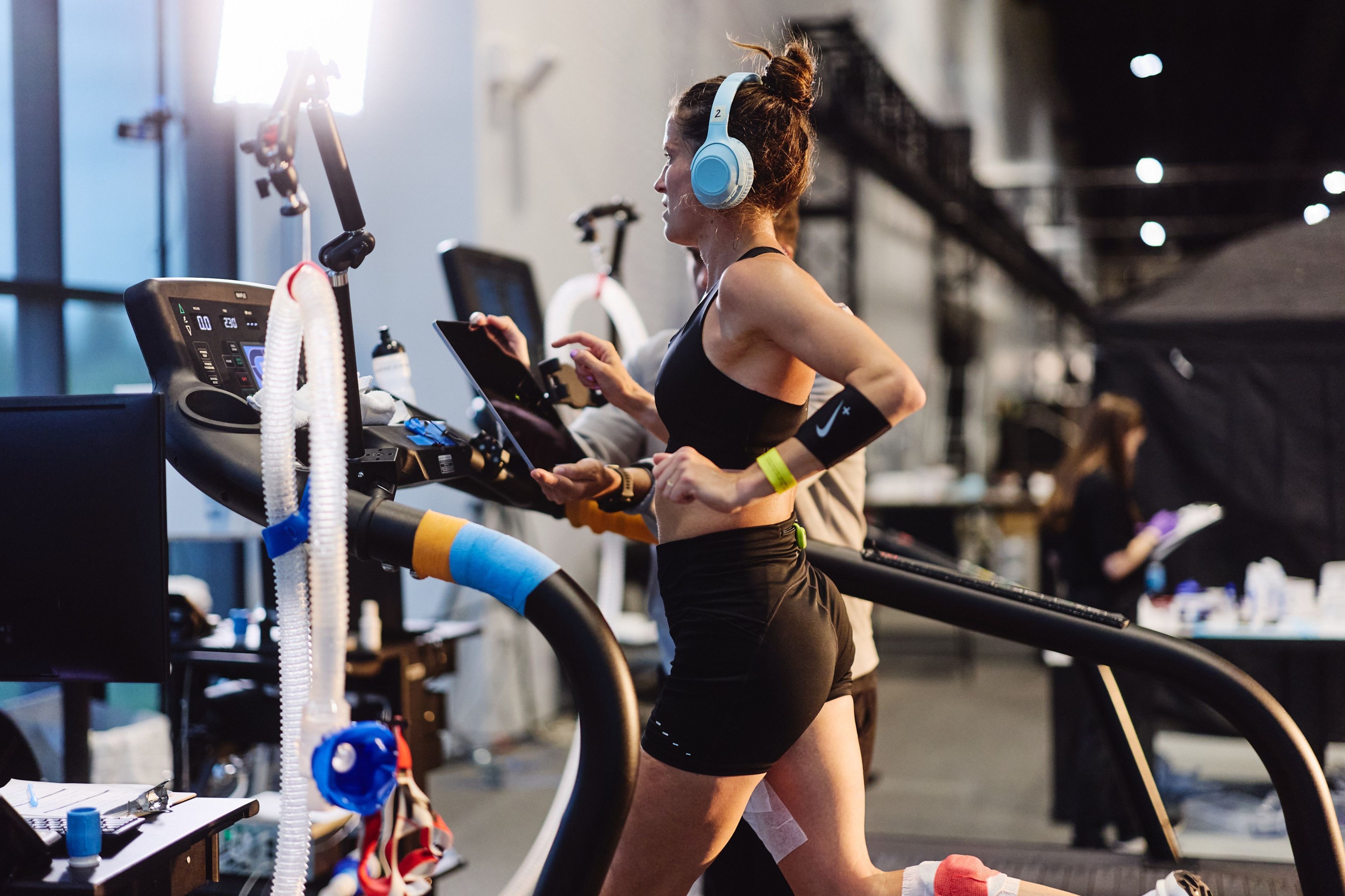 A woman wearing light blue headphones runs on a treadmill in the Nike Sport Research Lab, wearing black athletic wear with a Nike armband.