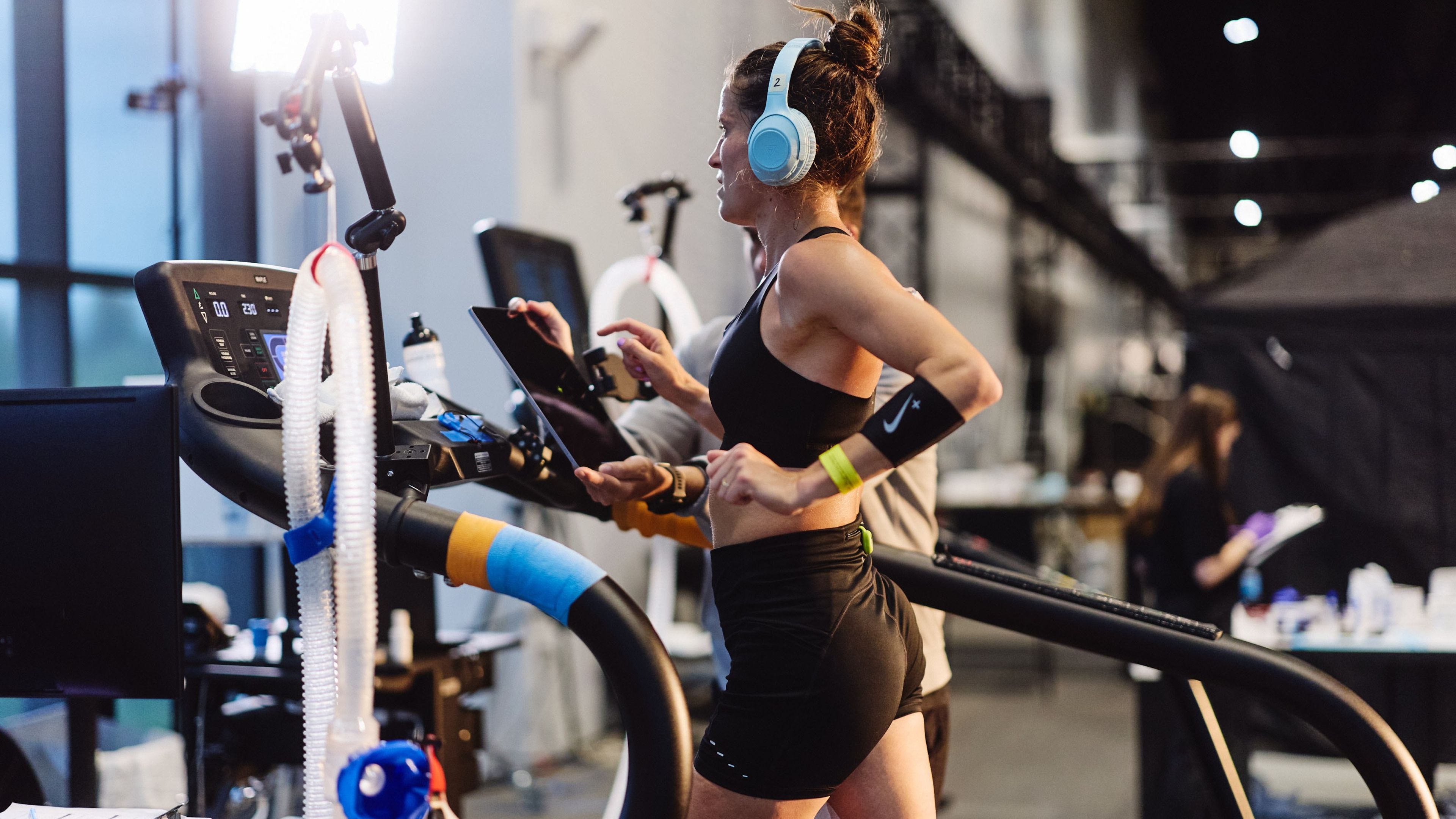 A woman wearing light blue headphones runs on a treadmill in the Nike Sport Research Lab, wearing black athletic wear with a Nike armband.