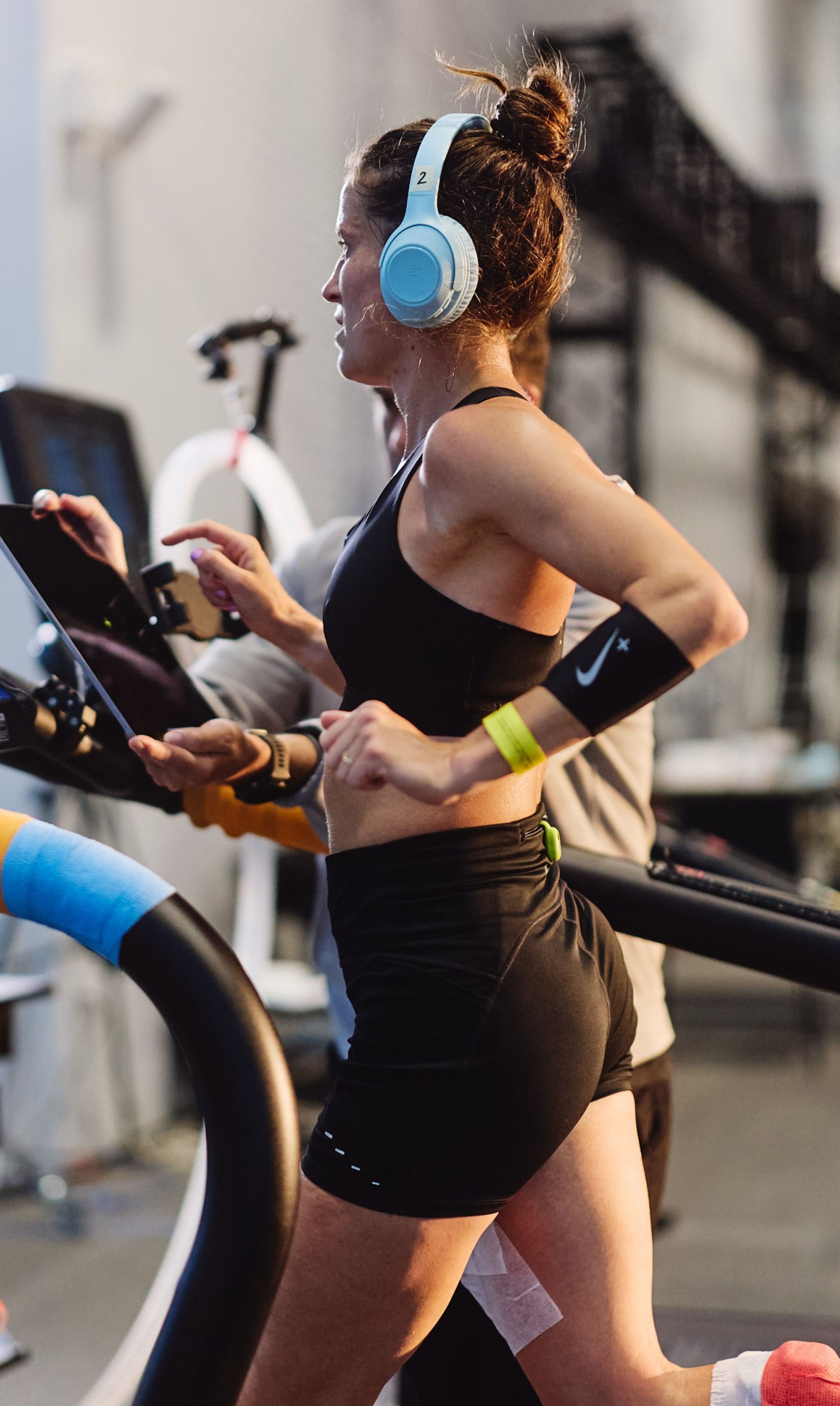 A woman wearing light blue headphones runs on a treadmill in the Nike Sport Research Lab, wearing black athletic wear with a Nike armband.