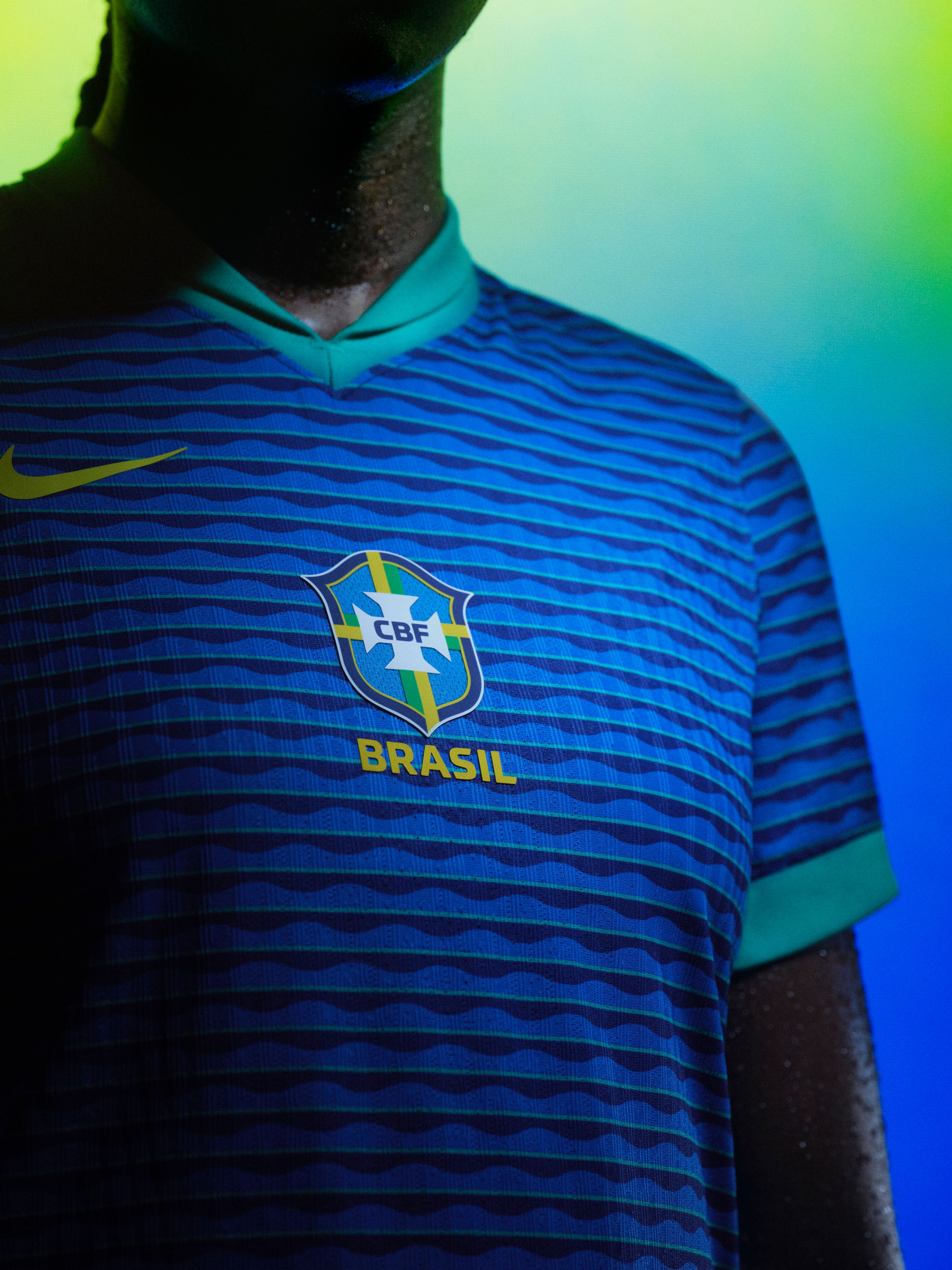 Close up view of an athlete's torso wearing a blue soccer jersey with teal trim on the cuffs and collar, featuring horizontal wave patterns, a yellow Nike swoosh on the right side of the chest, and a team logo in the center.