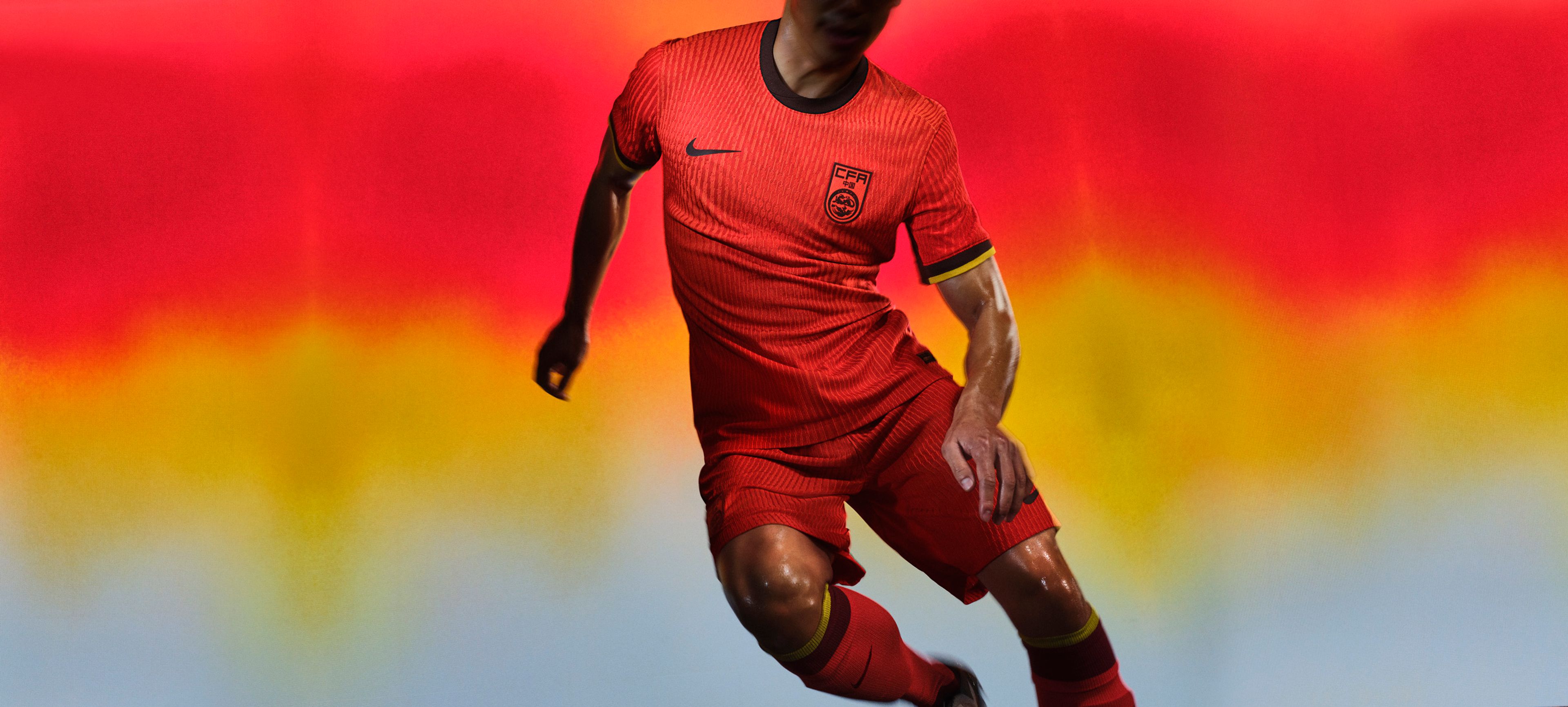 Athlete in a pivoting pose wearing a red-orange soccer jersey with black and yellow trim on the cuffs and a black collar, as well as matching red-orange shorts, each with a Nike swoosh and team logo on the chest and legs. They are also wearing socks with matching colors and trim.