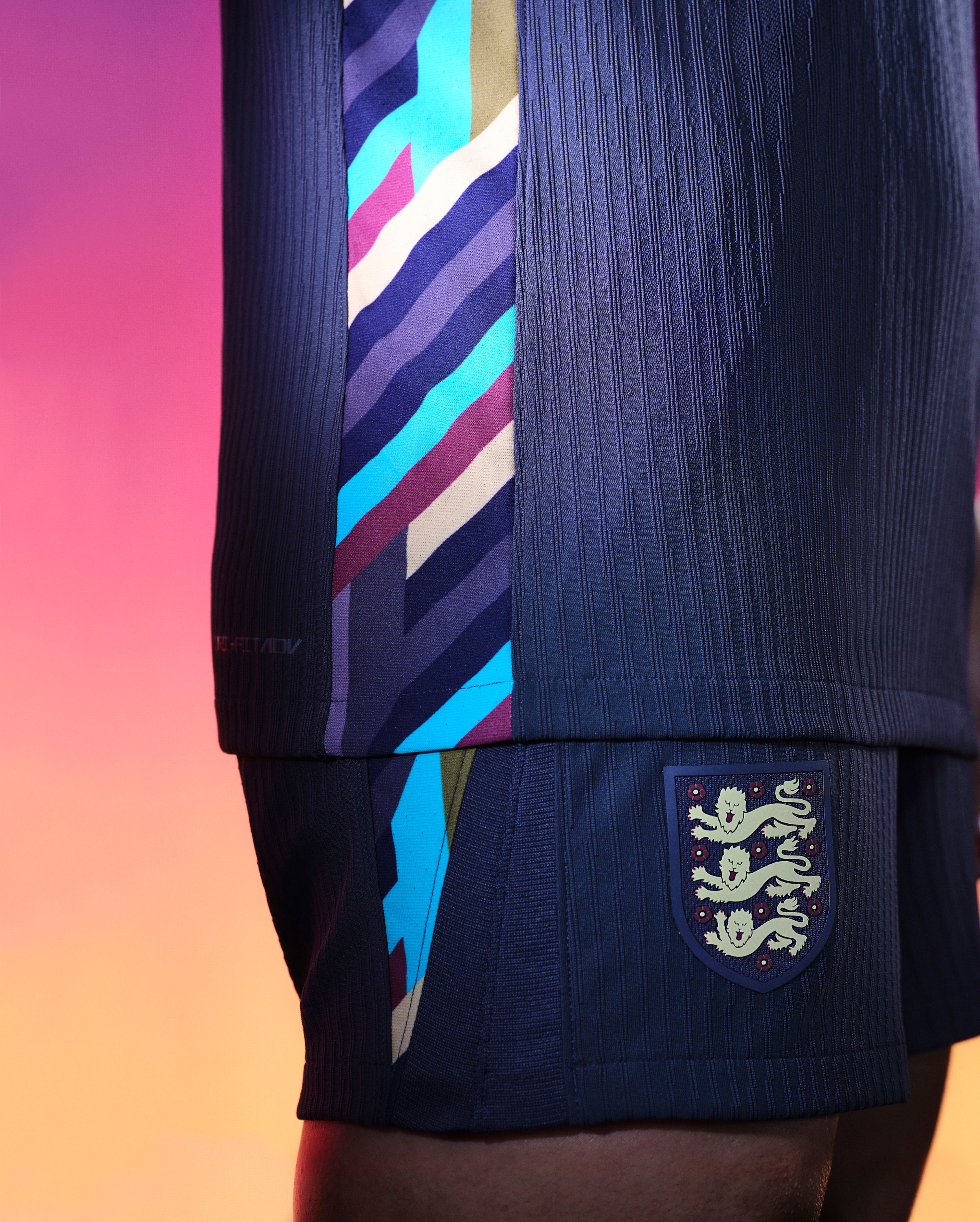 Close up side view of an athlete's lower torso wearing a navy blue soccer jersey and matching navy blue shorts. The jersey has a colorful diagonal stripe pattern on the side which continues and tapers off on the side of the shorts. There is a team logo on the right pant leg.