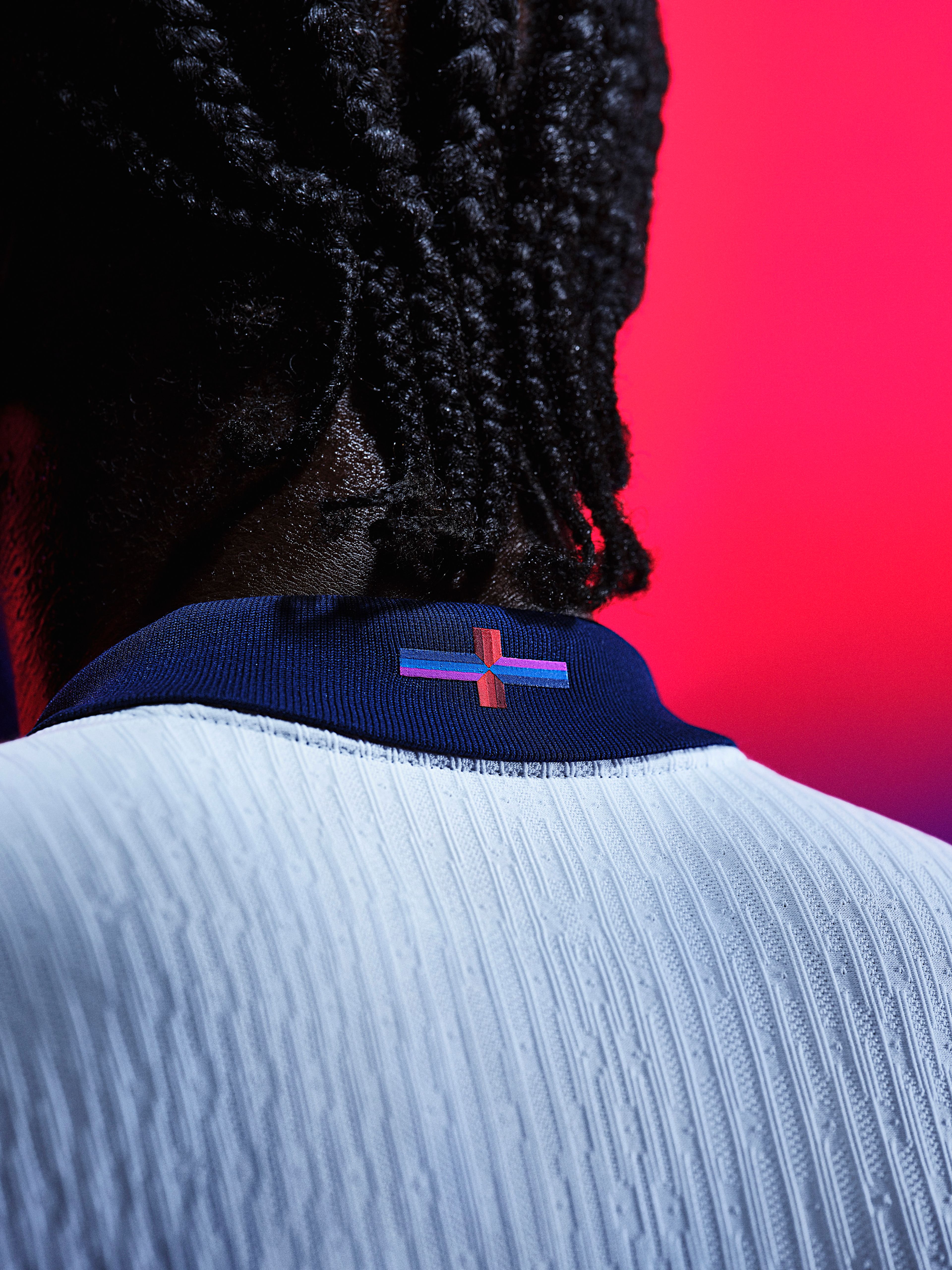 Close up back angle view of an athlete wearing a white soccer jersey that has a dark blue collar with a logo in the rear center.