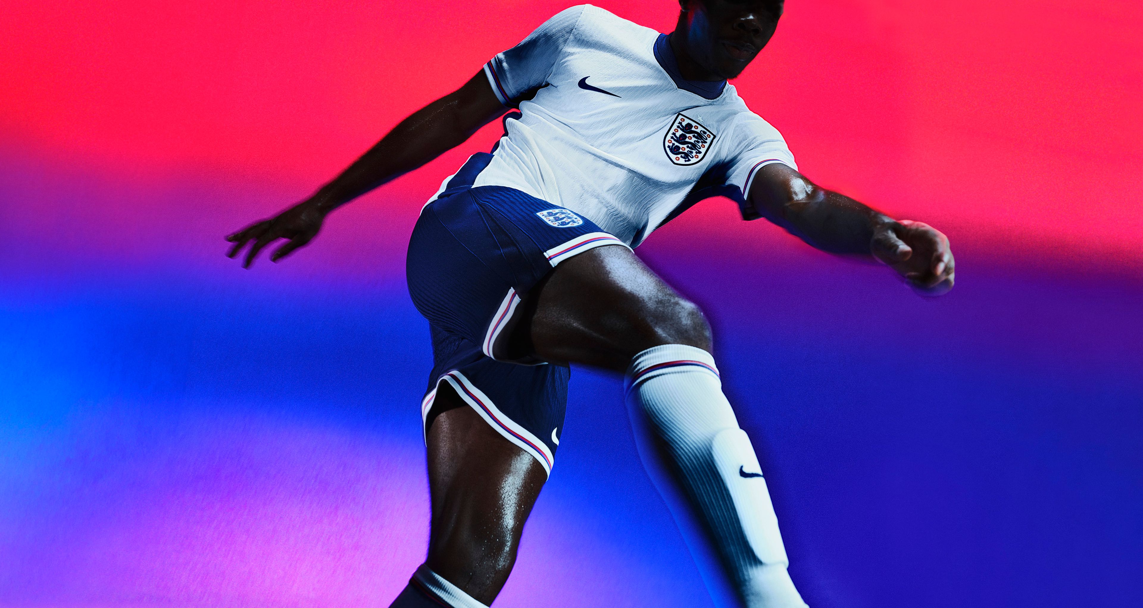 Athlete in a kicking pose wearing a white soccer jersey with a dark blue collar, dark blue sides, and red, white, and blue striped on the cuffs, as well as indigo blue shorts with matching trim, each with a Nike swoosh and team logo on the chest and legs. They are also wearing white socks with matching trim.