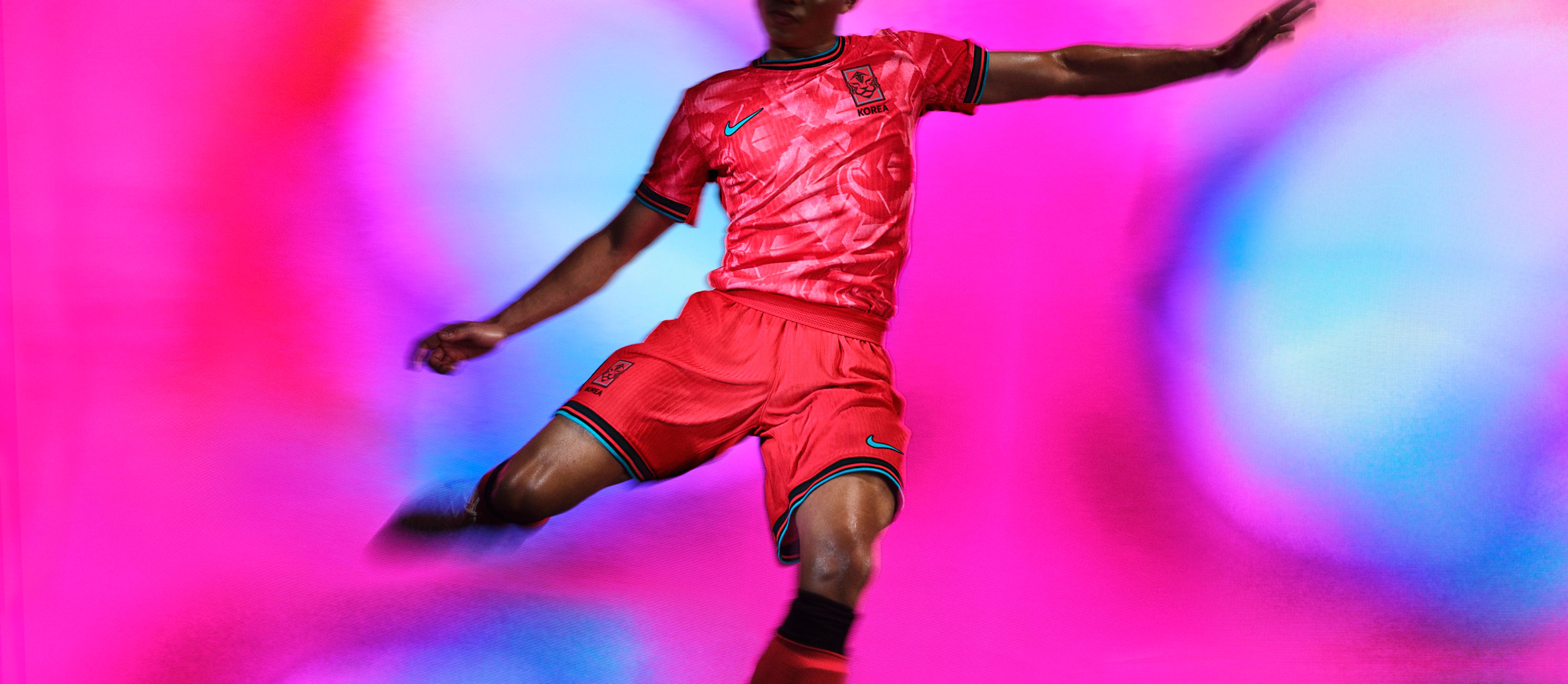 Athlete in a mid-kicking pose wearing a red and pink soccer jersey with black and teal striped trim, as well as and matching red shorts with matching trim, each with a Nike swoosh and team logo on the chest and legs.