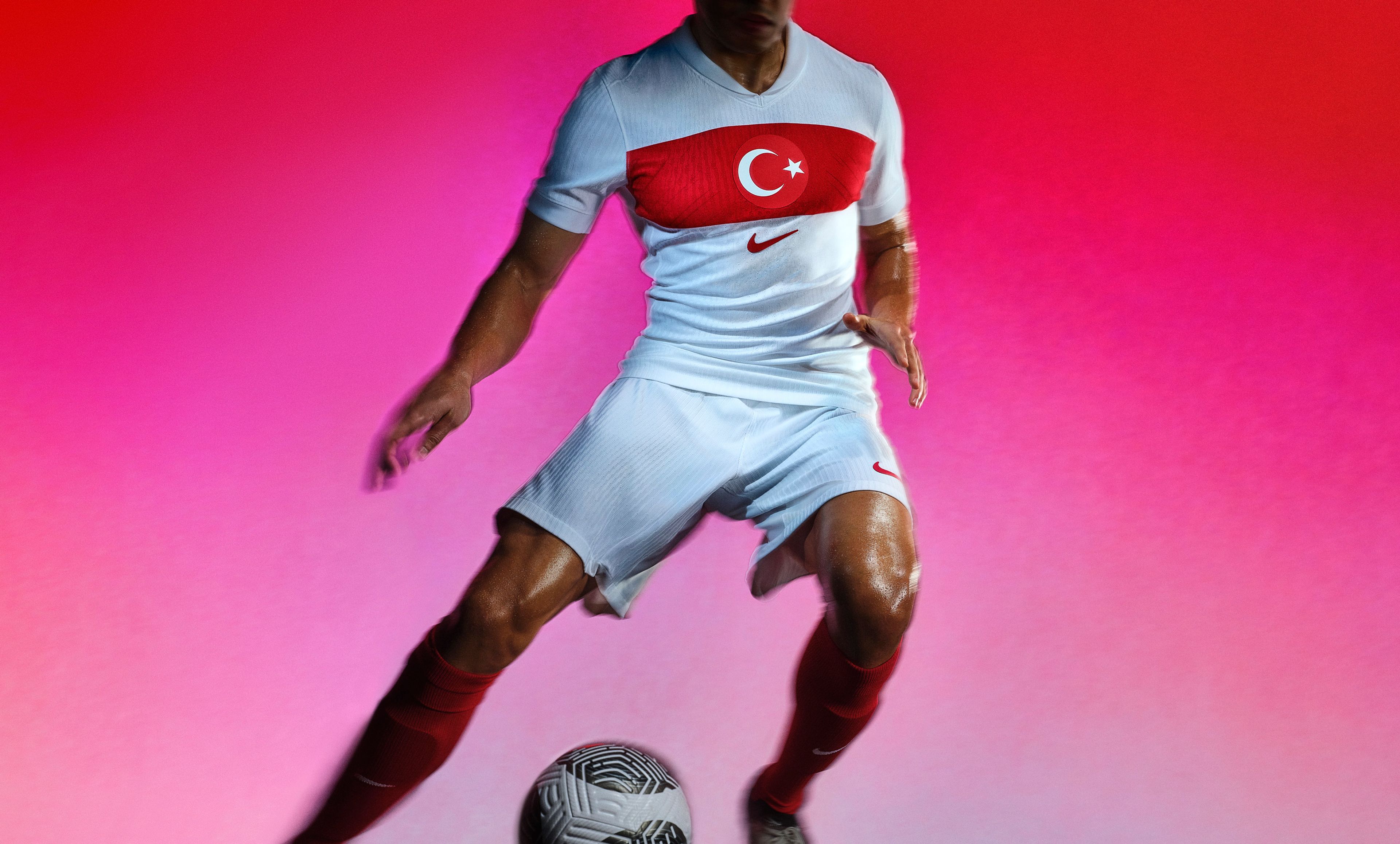 Athlete dribbling a soccer ball wearing a white soccer jersey with a red stripe across the chest, as well as matching white shorts, each with a Nike swoosh on the chest and legs.