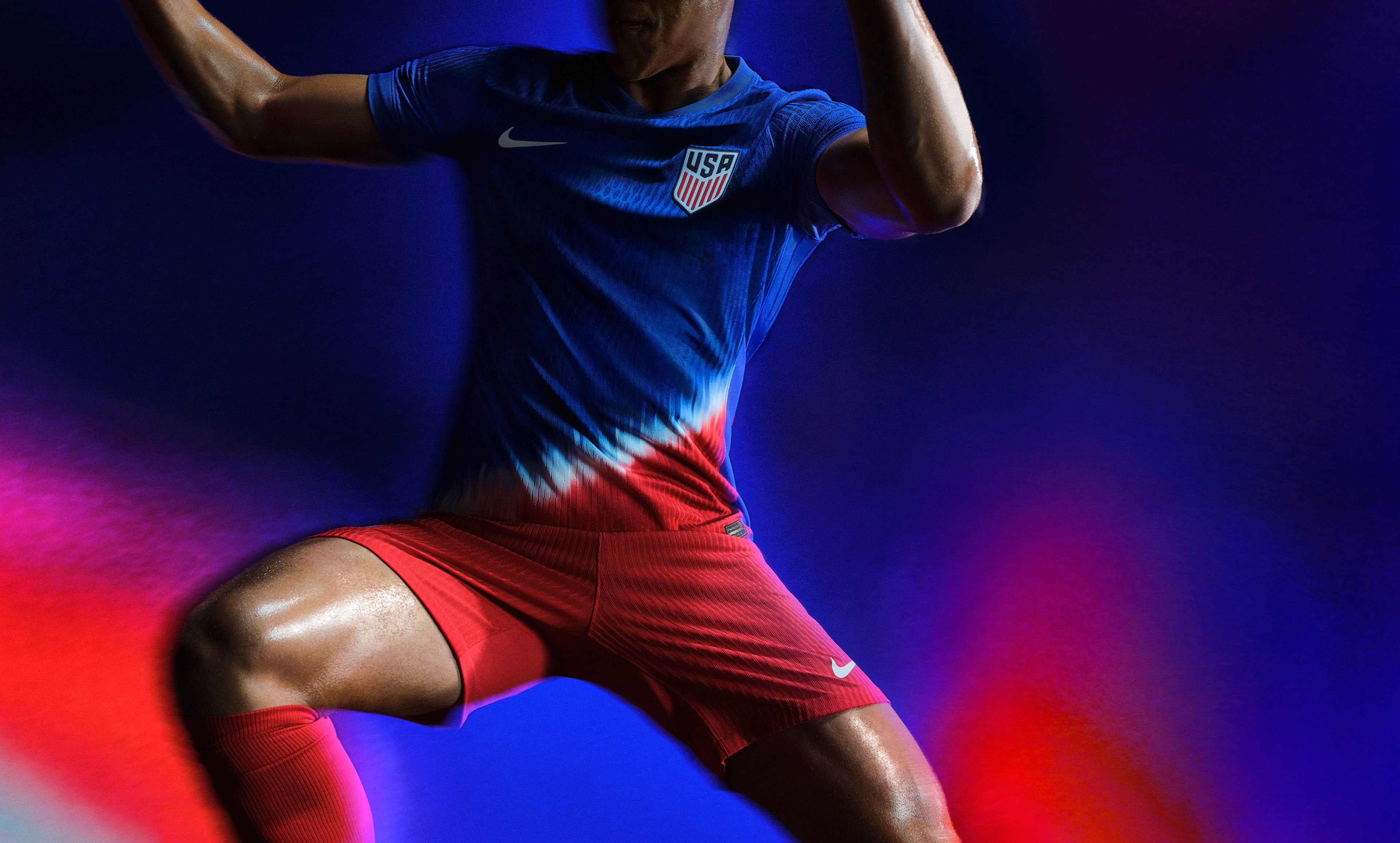 Athlete in a mid-kicking pose wearing a blue soccer jersey with a white and red diagonal stripe on the stomach, as well as matching red shorts, each with a Nike swoosh on the chest and legs and a team logo on the chest.