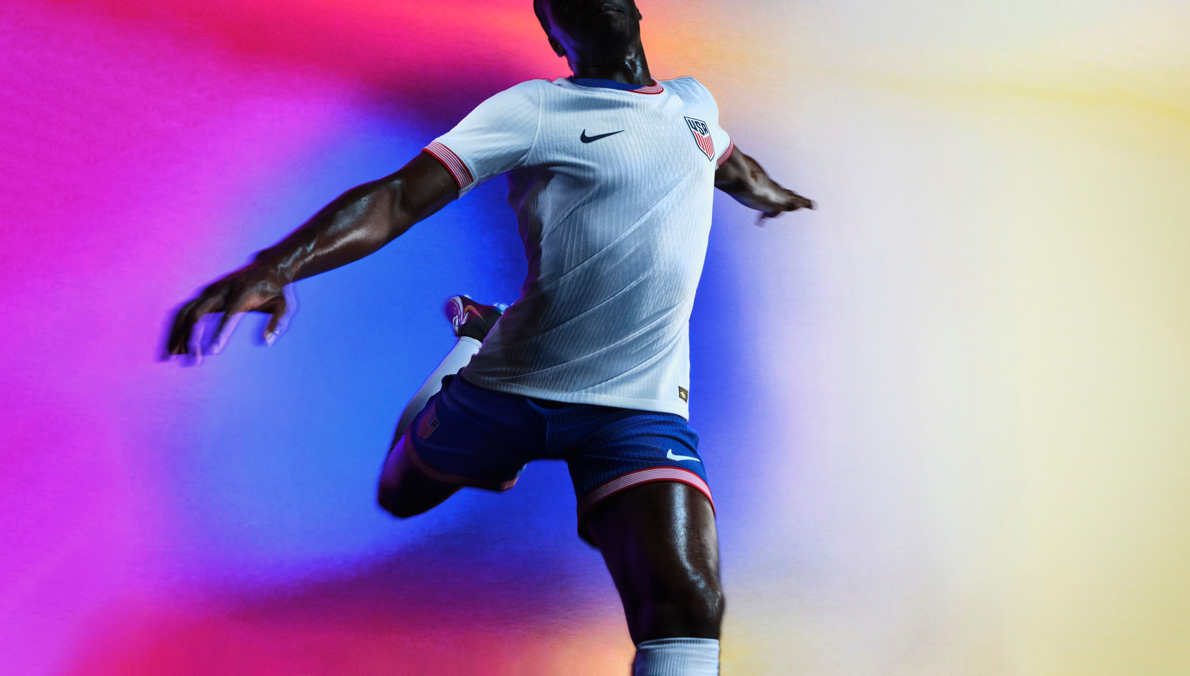 Athlete in a mid-kicking pose wearing a white soccer jersey with red and white striped trim on the collar and cuffs, as well as blue shorts with red and white striped trim, each with a Nike swoosh and team logo on the chest and legs.