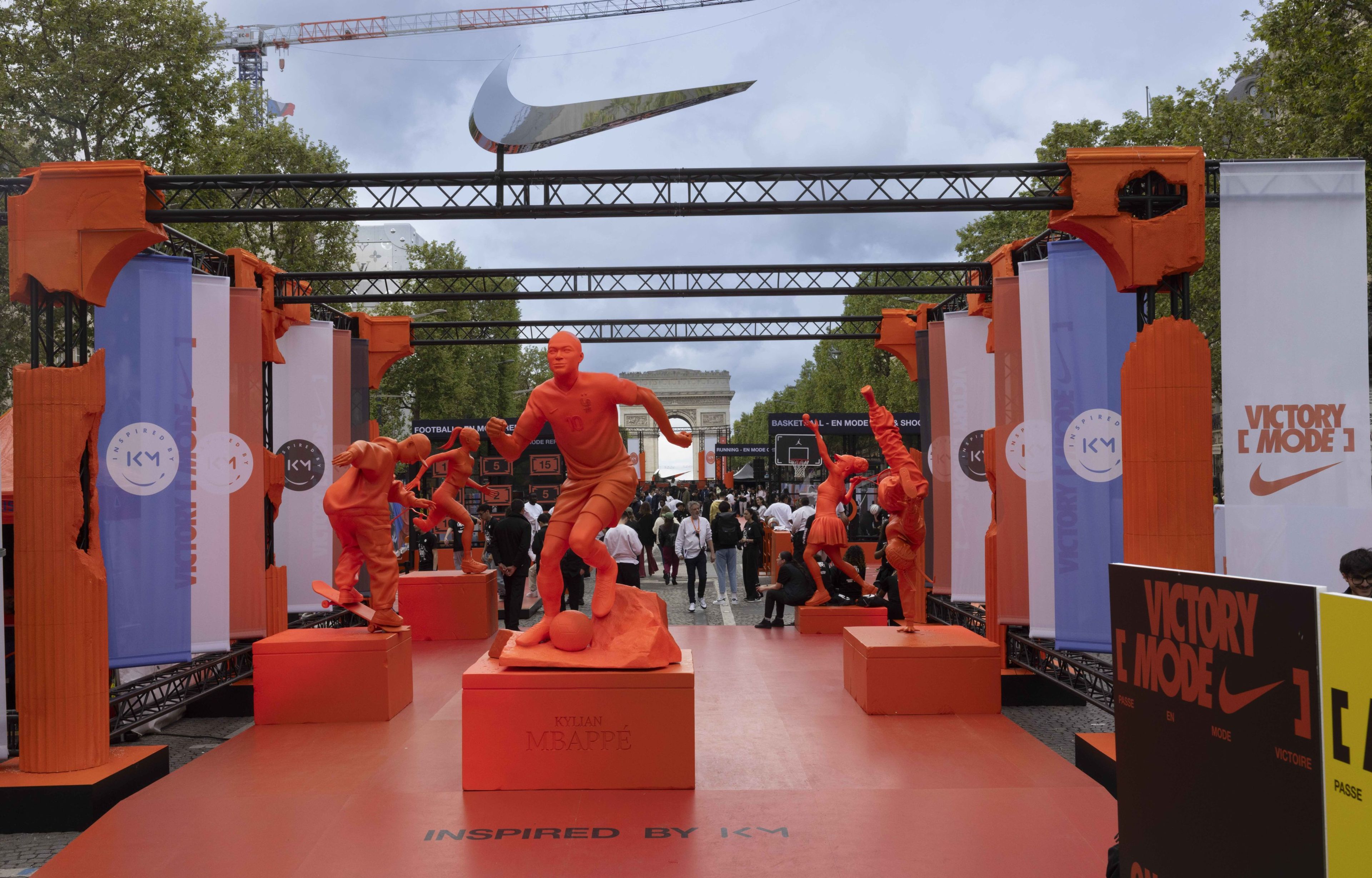 Front view of a pavilion of bright orange statues depicting athletes doing different sports, at the Victory Mode event in Paris, France. Statues are surrounded by orange pillars and blue, white, and orange banners.