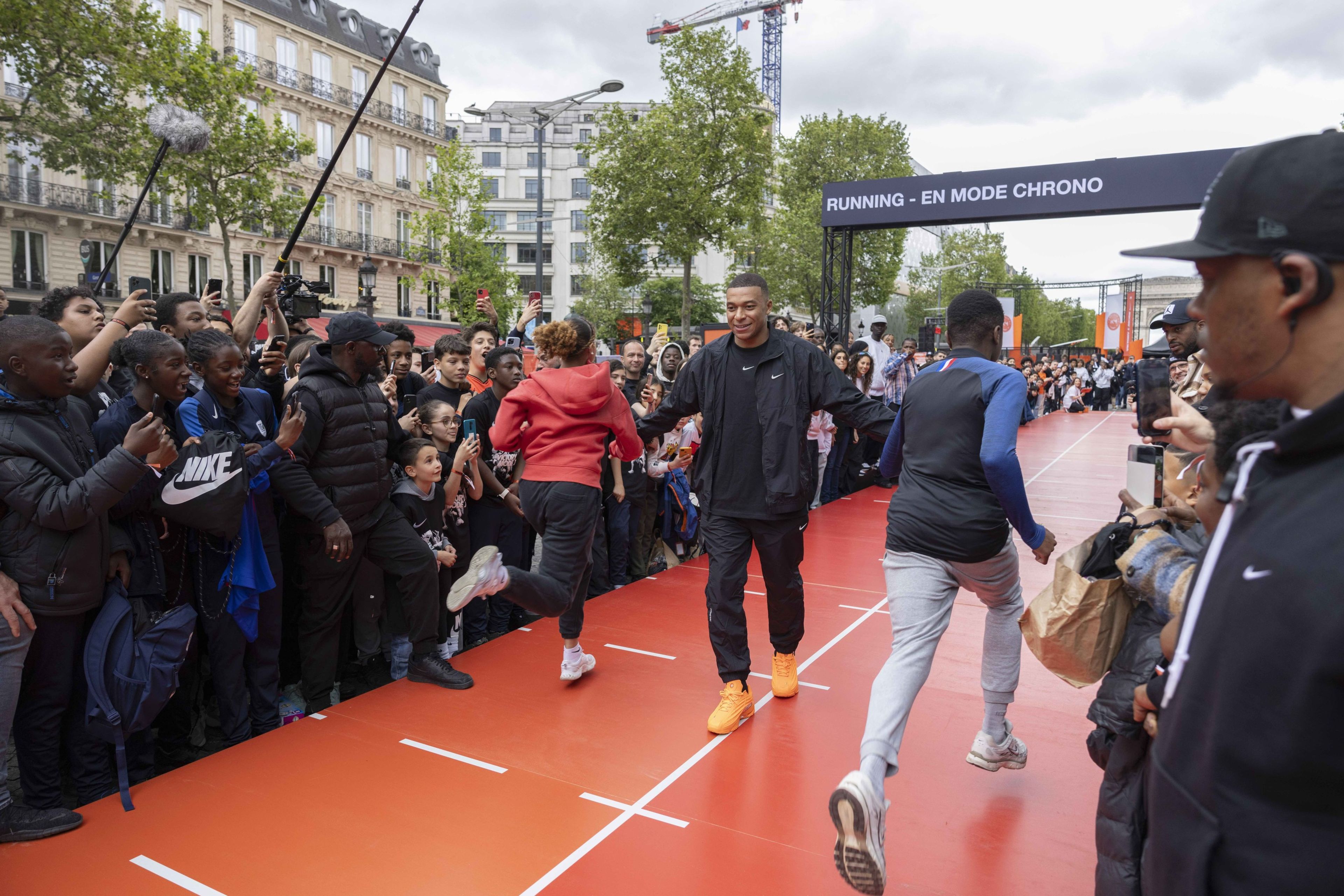 Front view of Kylian Mbappe wearing a black Nike tee, jacket, and pants, while walking between 2 runners on an orange stage at the Victory Mode event in Paris, France. A crowd surrounds the activity on the stage.