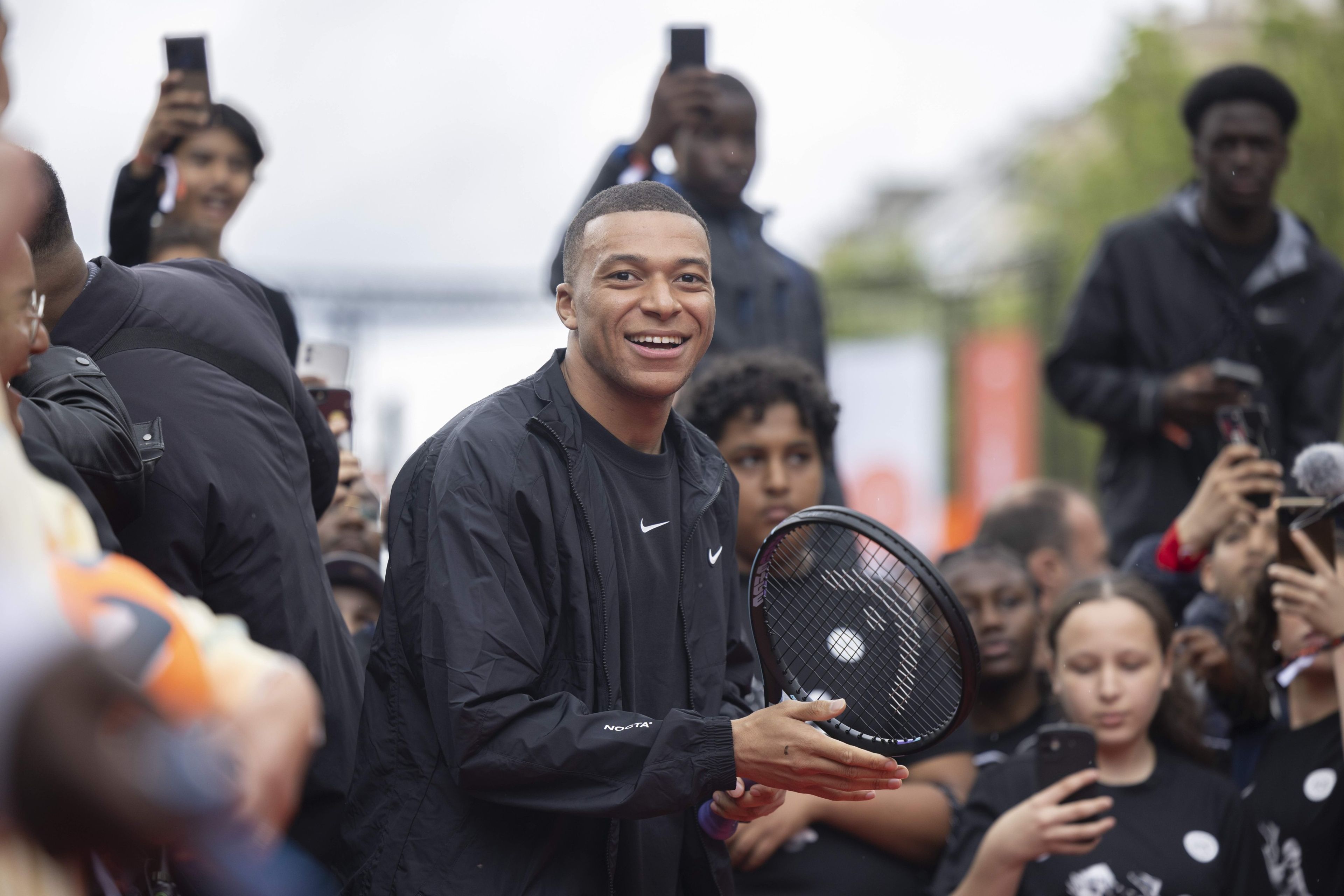 Front view of Kylian Mbappe, wearing a black Nike tee and jacket, smiling as he holds a tennis racket in front of a crowd of kids at the Victory Mode event in front of the Nike House of Innovation in Paris, France.
