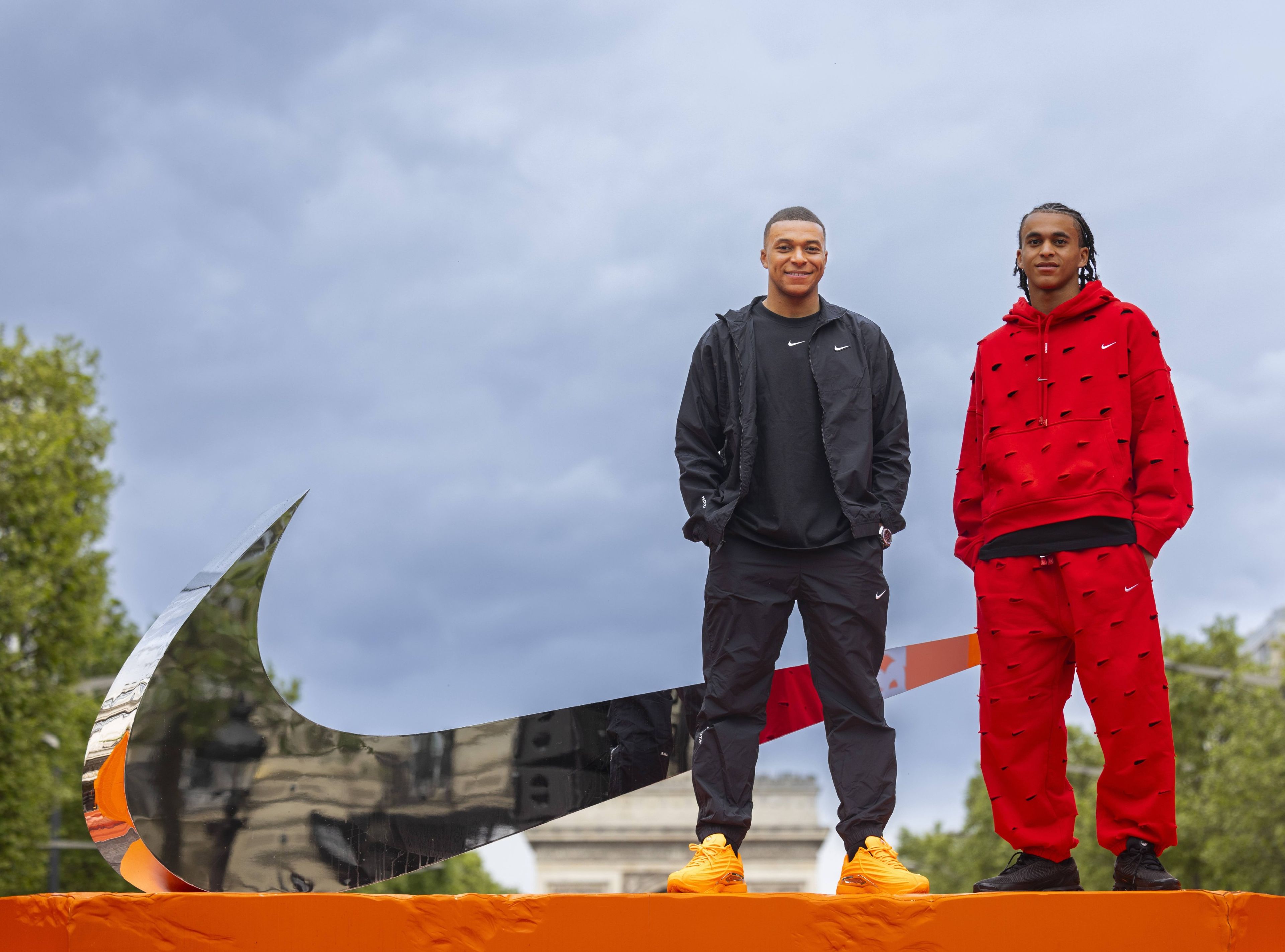 Front view of Kylian Mbappe, and his brother, Ethan Mbappe, as they stand next to a large mirrored Swoosh. Kylian wears black Nike tee, jacket and pants with orange footwear. Ethan wears a red fleece hoodie and pants with black footwear. Both smile towards the camera.