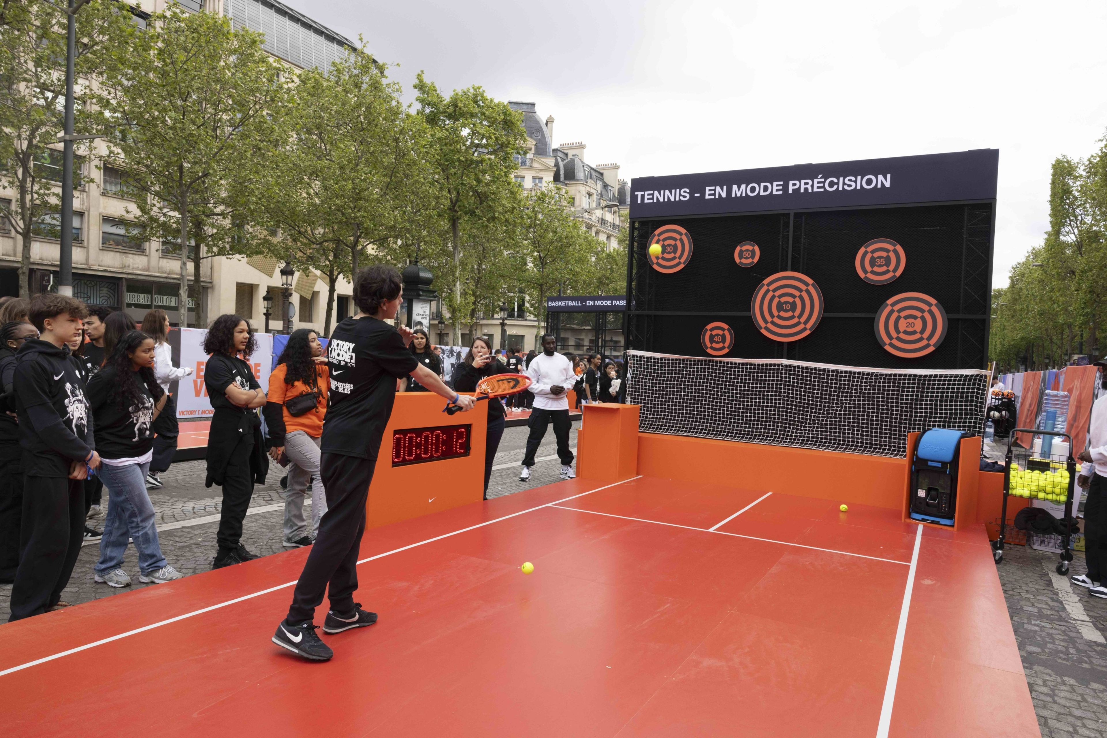 Back view of a young athlete wearing a black Victory Mode tee using a bright orange tennis racket to participate in the Tennis Challenge at the Victory Mode event in Paris, France. A crowd looks on as he aims at orange and black targets.