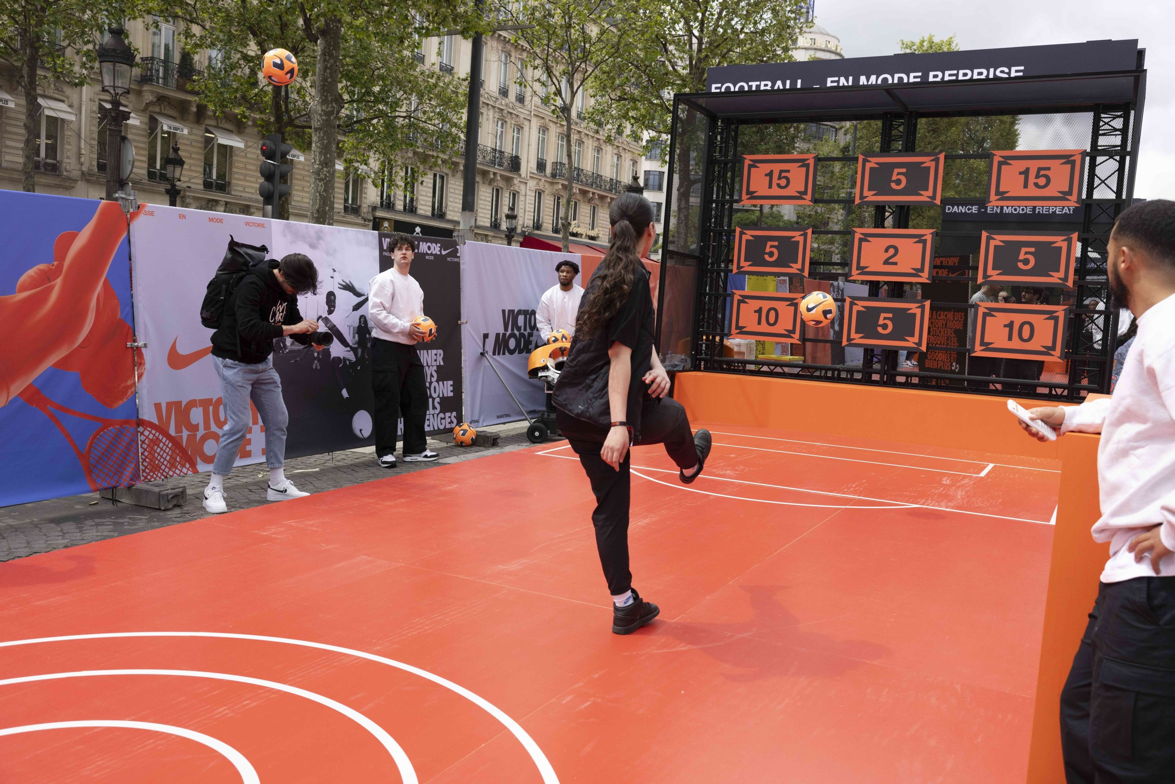 Back view of a young athlete kick an orange football during a football challenge at the Victory Mode event in Paris, France. She stands on a bright orange court, with photographers and event helpers nearby.