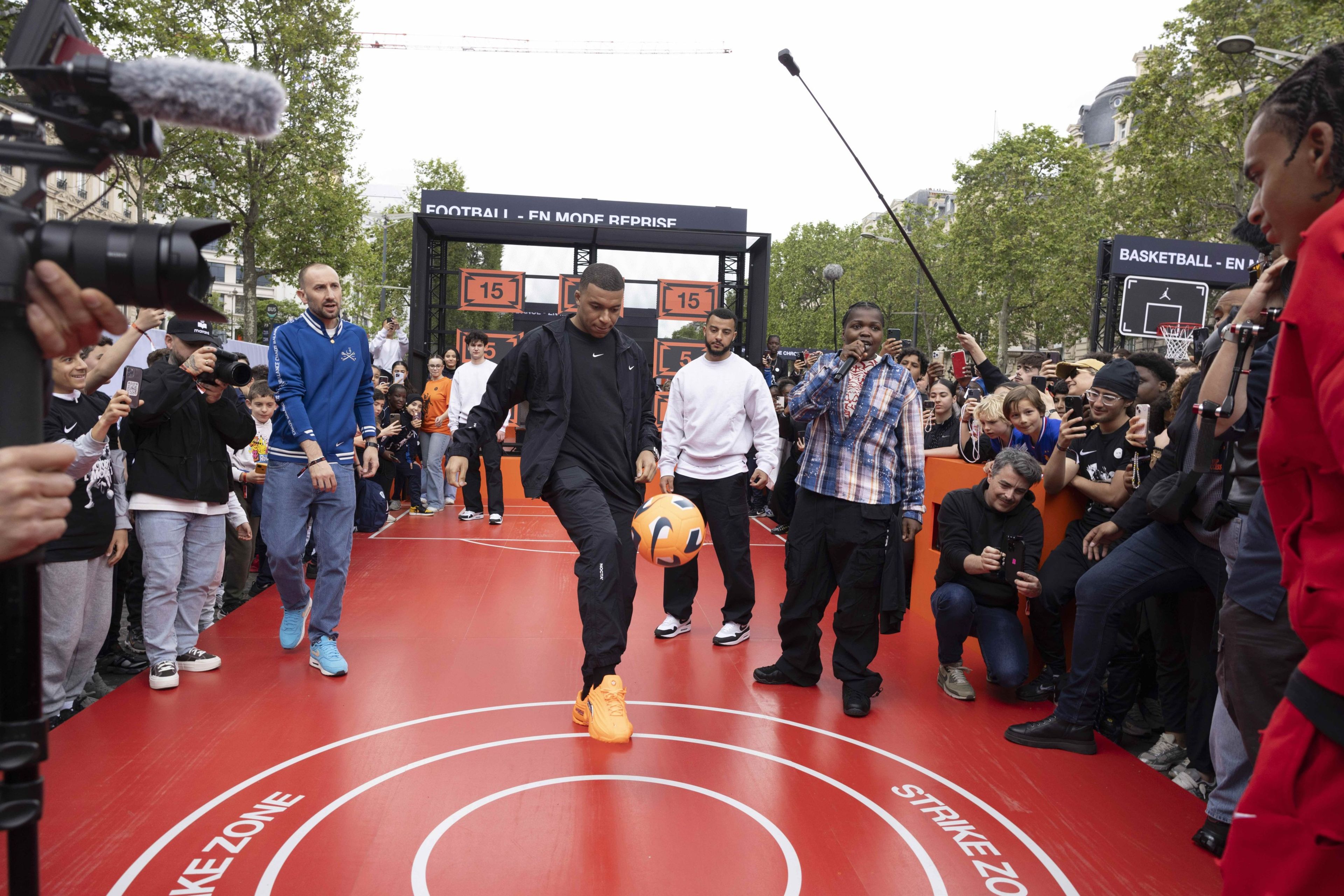 Front view of Kylian Mbappe juggling an orange Nike football on a red platform in front of a crowd of young athletes and photographers at the Victory Mode event in Paris, France.