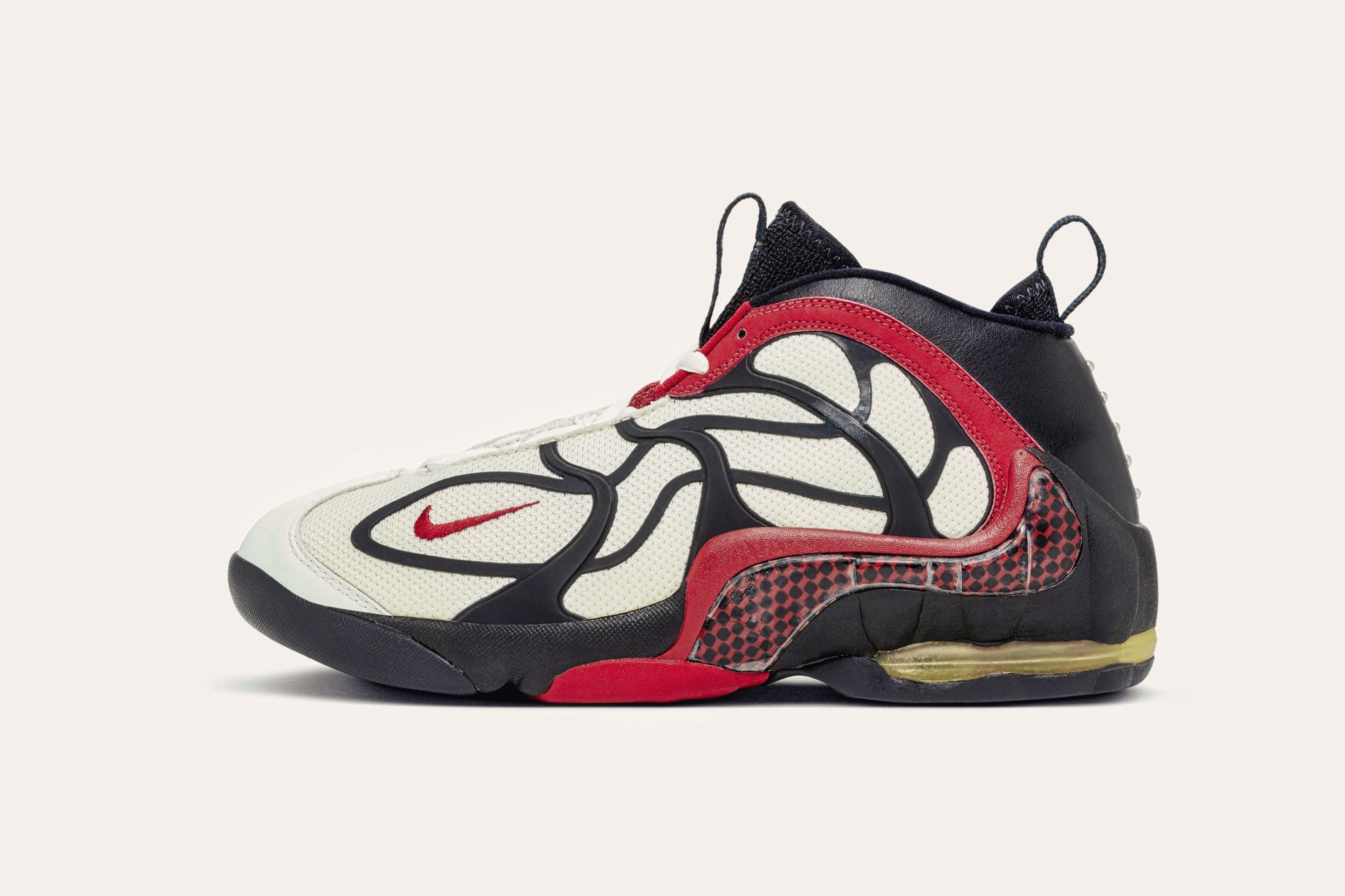 A white sneaker with a black leather backing and a black web-like pattern with red trim.