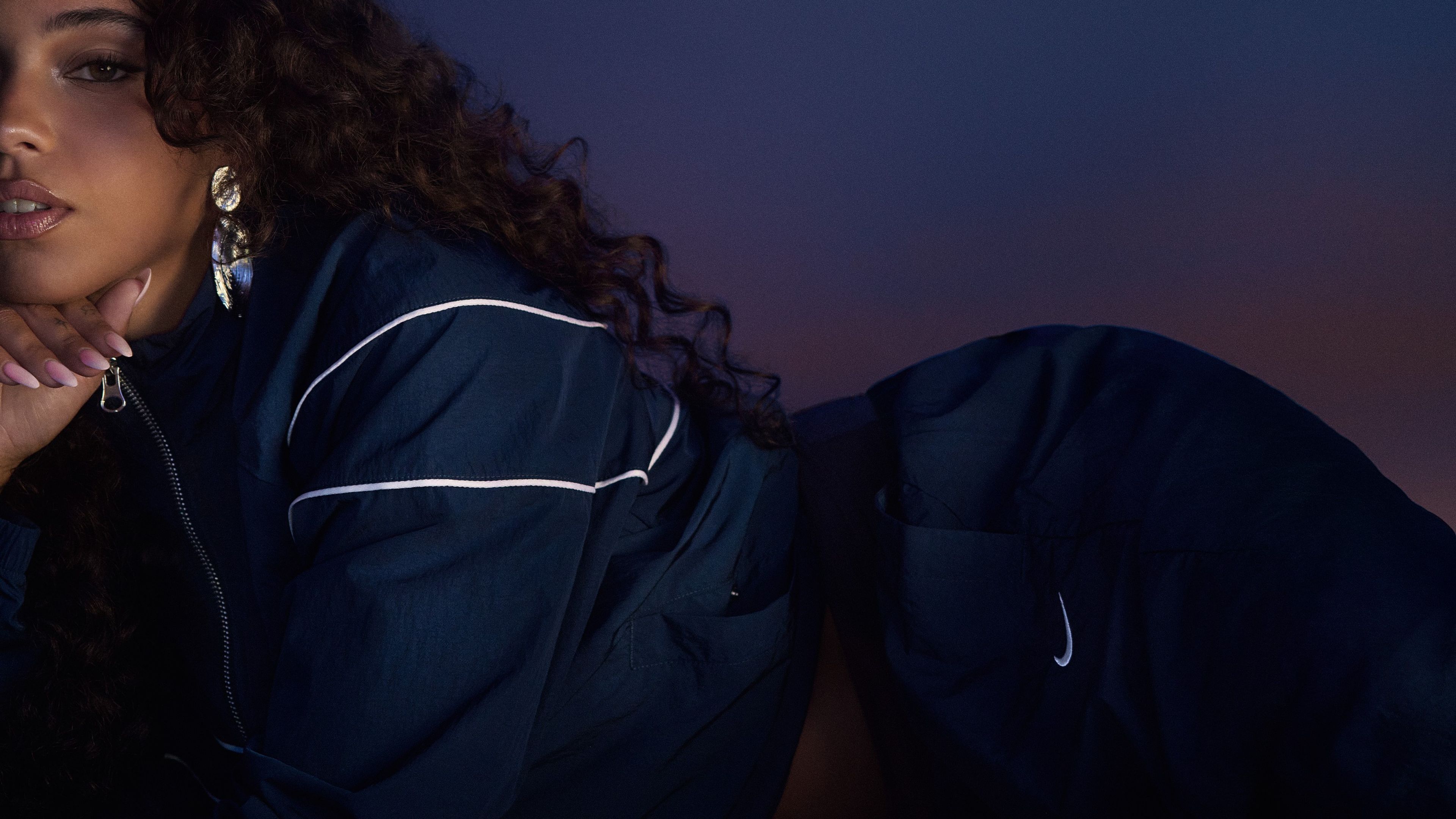 Maria Isabel lies against a blue/purple gradient background, wearing a navy blue Nike Windrunner tracksuit. We see part of her body and face, as she props her hand under her chin, and looks directly at the camera. She wears silver earrings that reflect the blue/purple light.
