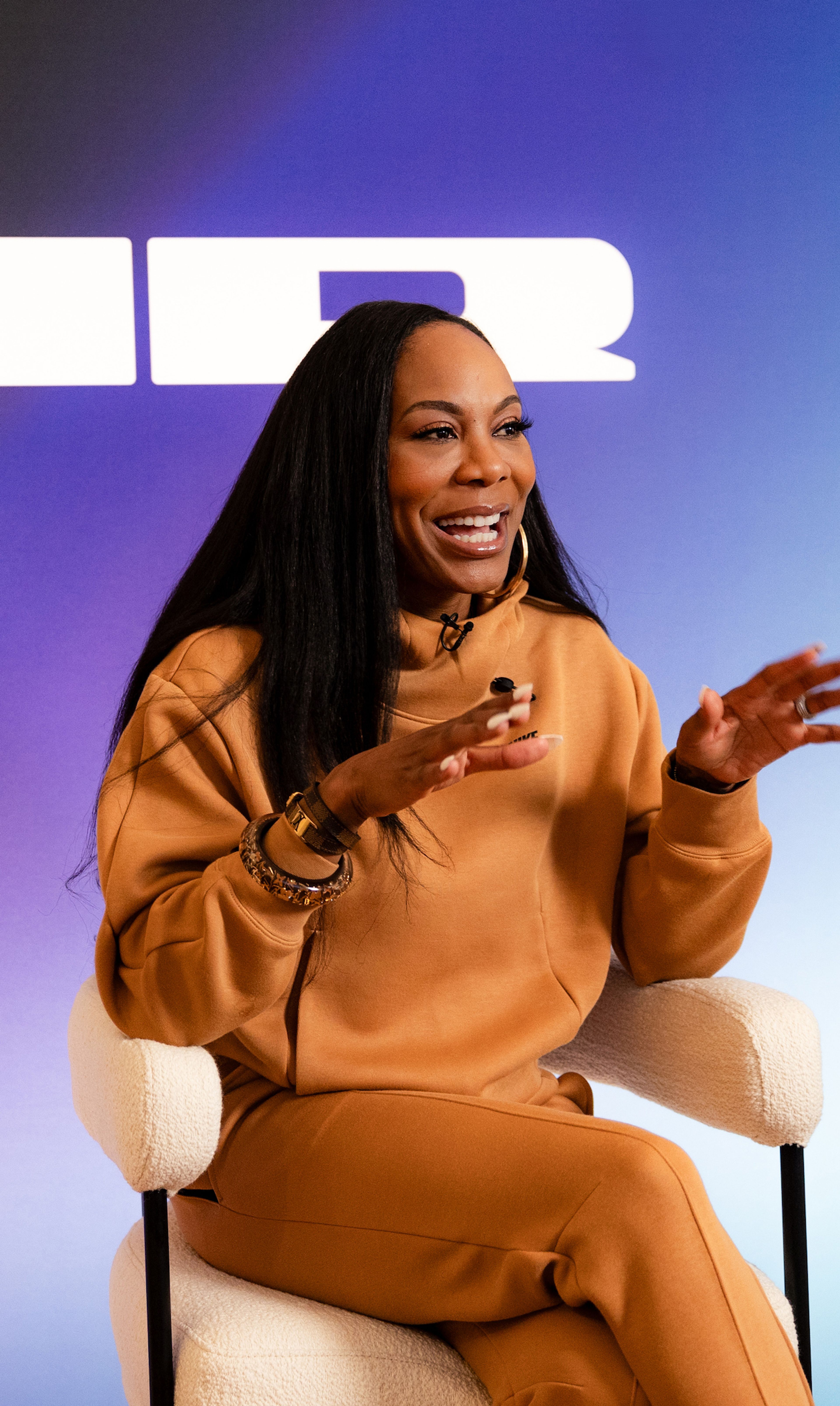 Front angled view of Sanya Richards-Ross wearing Nike Sportswear Tech Fleece in the Flax color. Sanya sits on a white stool, against a blue & purple gradient background with a large white logo for Nike AIR.