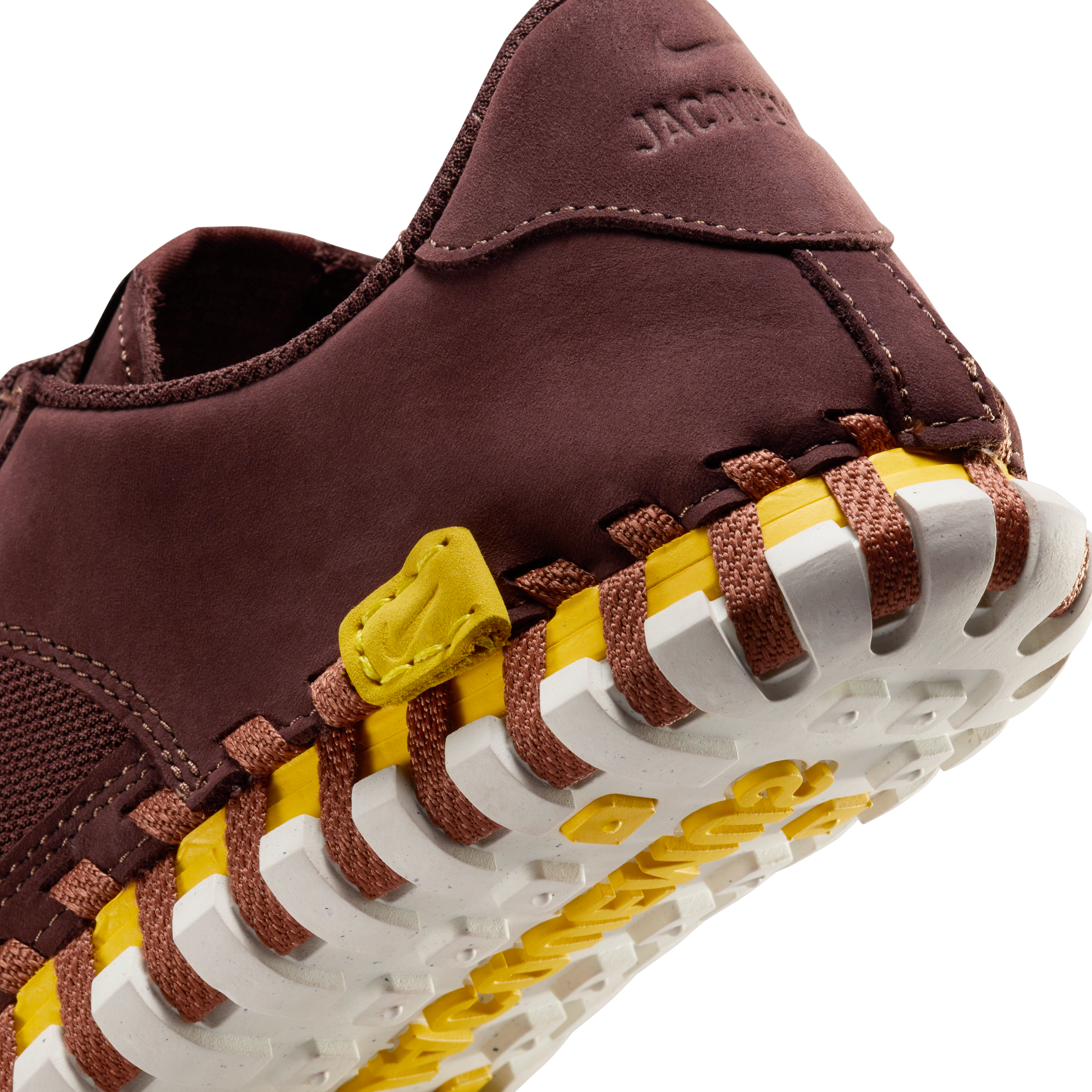 Back detail view of a Nike x Jacquemus J Force 1 Low LX SP with Earth brown upper, tour yellow midsole, and white sole. There is a small sewn-in yellow leather label at the base of the heel with an embossed Swoosh, and an embossed Jacquemus logo on the heel cup. The shoe floats on a black background.