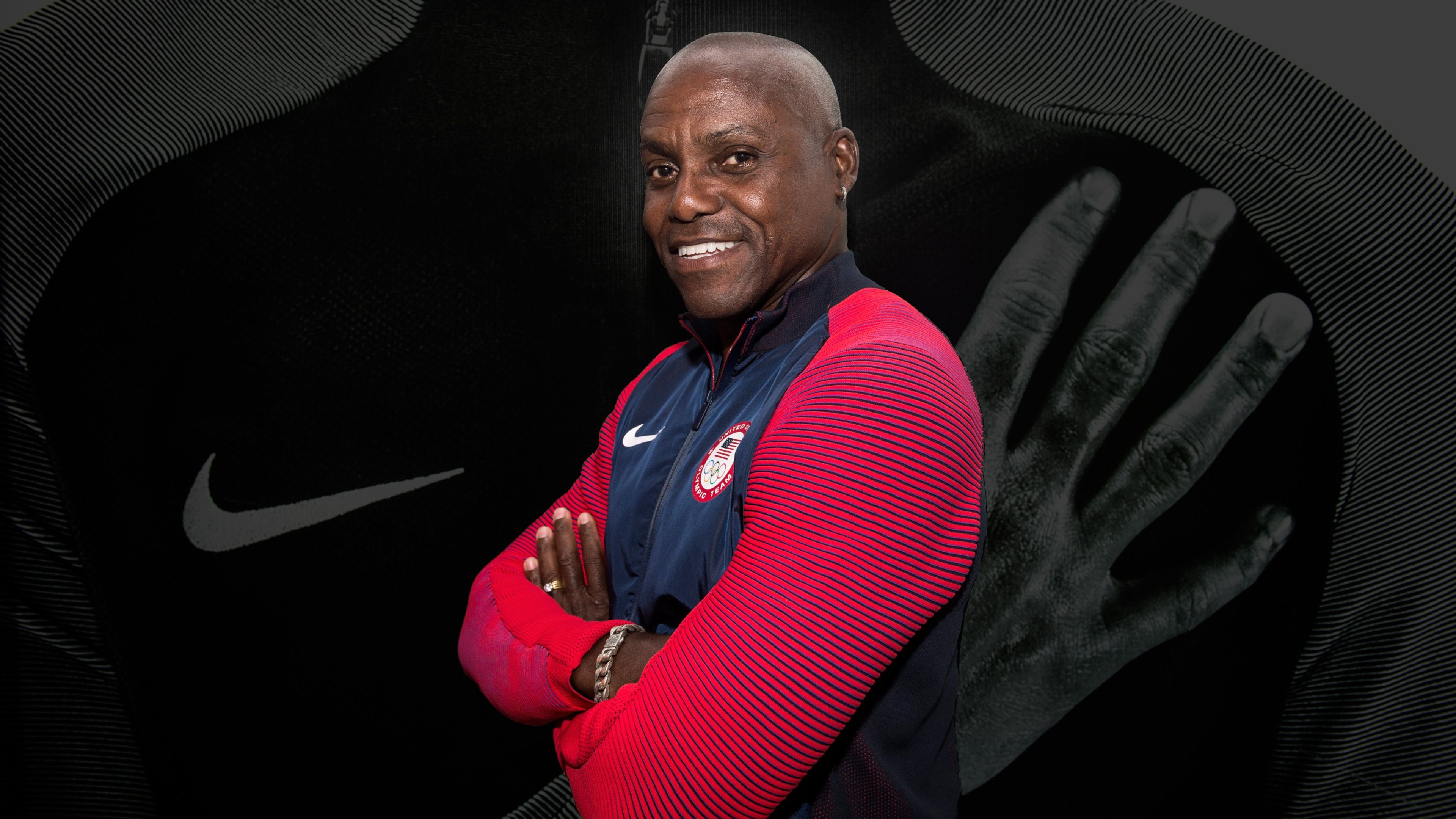 Carl Lewis wearing a red and blue Nike olympics jacket, smiling with his arms folded. In the background is a dimmed photo of his torso with his hand over his heart.