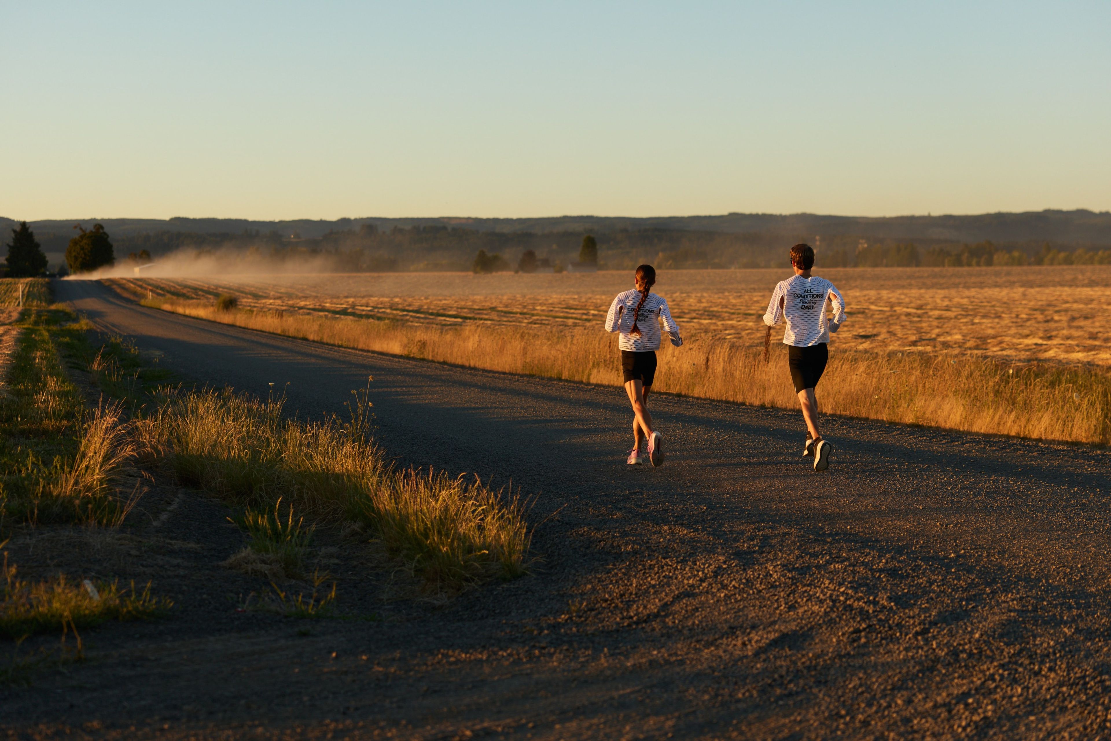 Two runners in Nike Radical Airflow tops jog down a gravel road through golden farmland at sunrise.