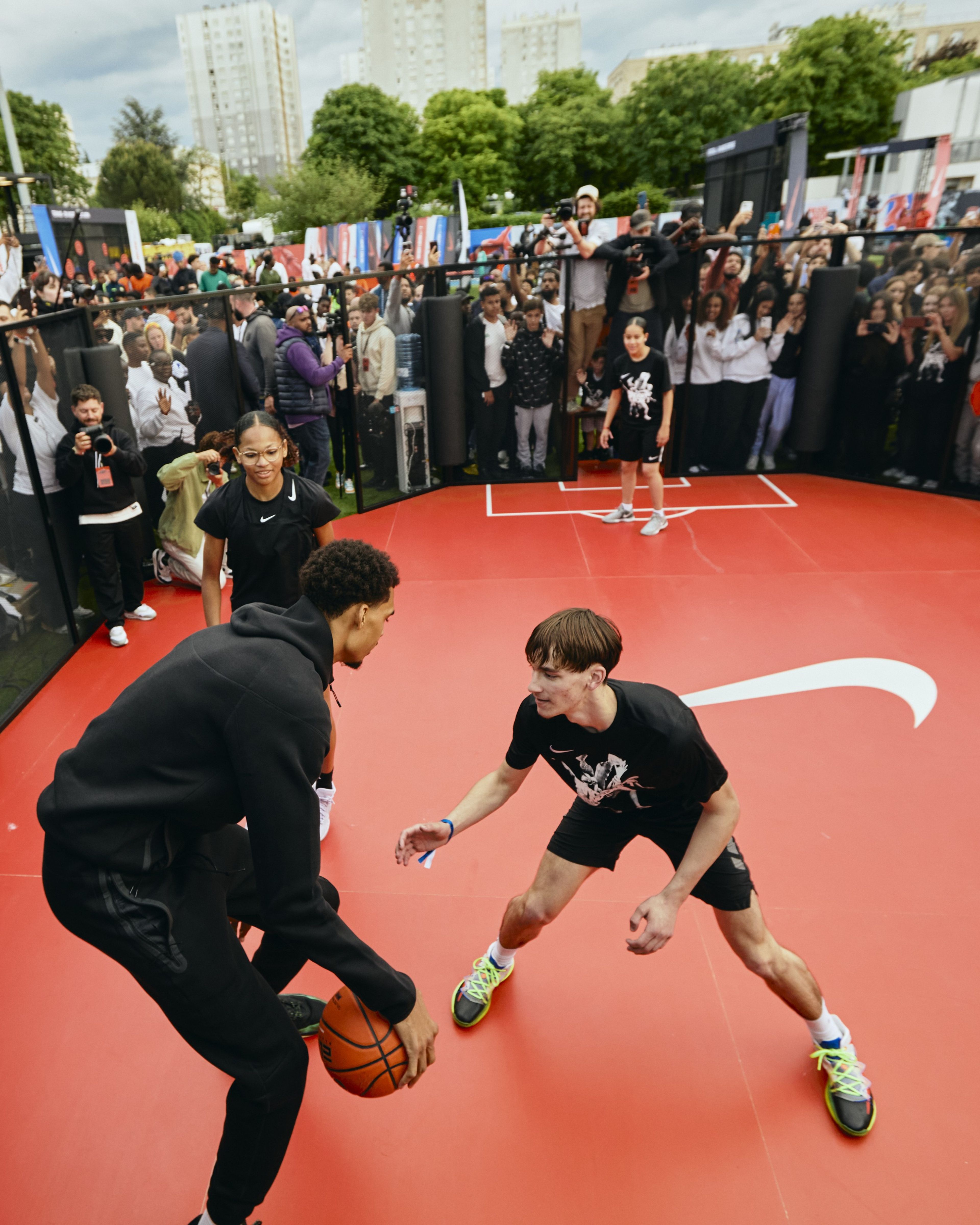 Victor Wembanyama plays one on one basketball with a young athlete, as a crowd behind a fence cheers them on. They play on a red court displaying a white Nike Swoosh, and two other athletes wait to join the game.