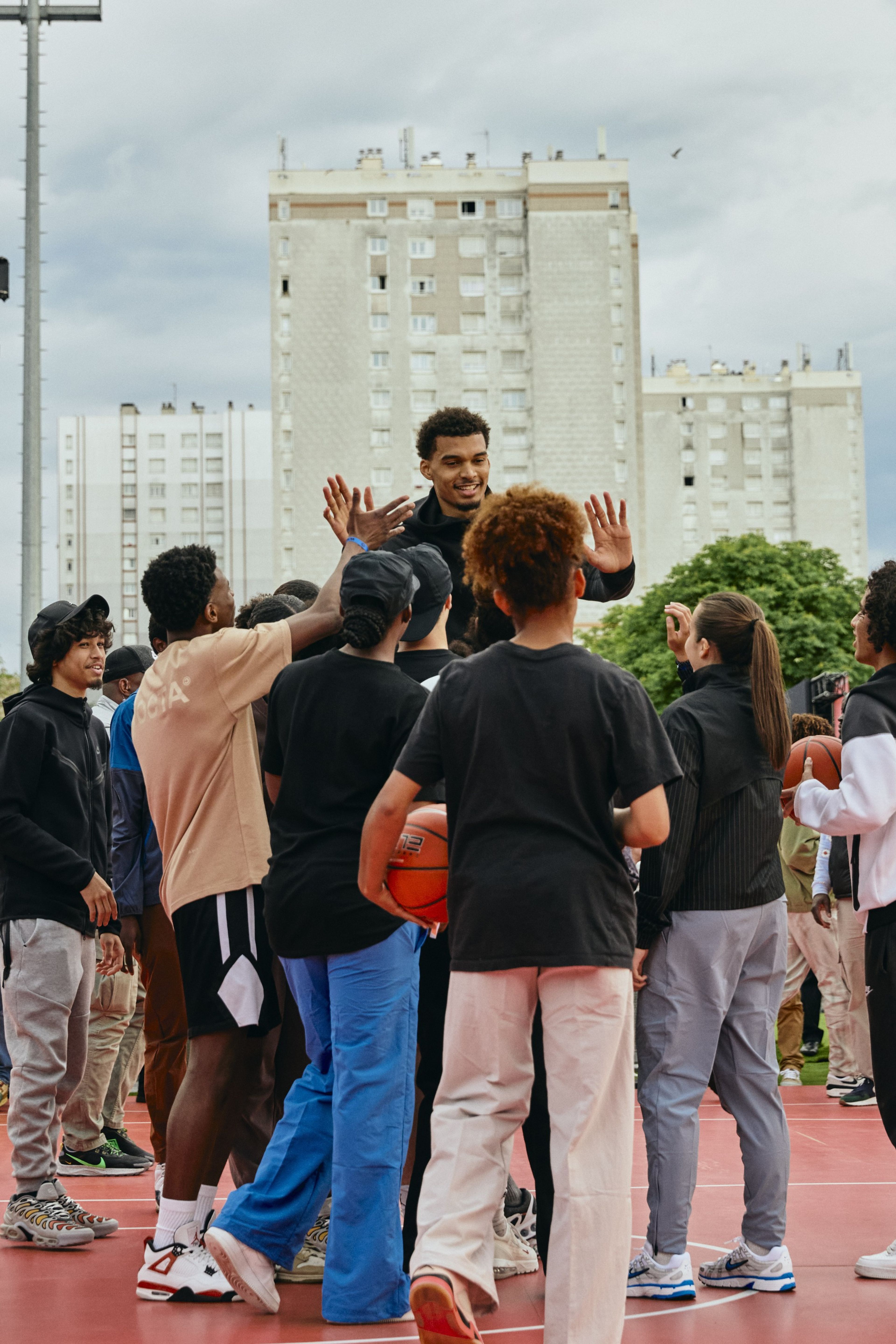Victor Wembanyama gives high fives to a crowd of young athletes at the Victory Mode event in Nanterre, France. They stand on a red court with white buildings in the background.