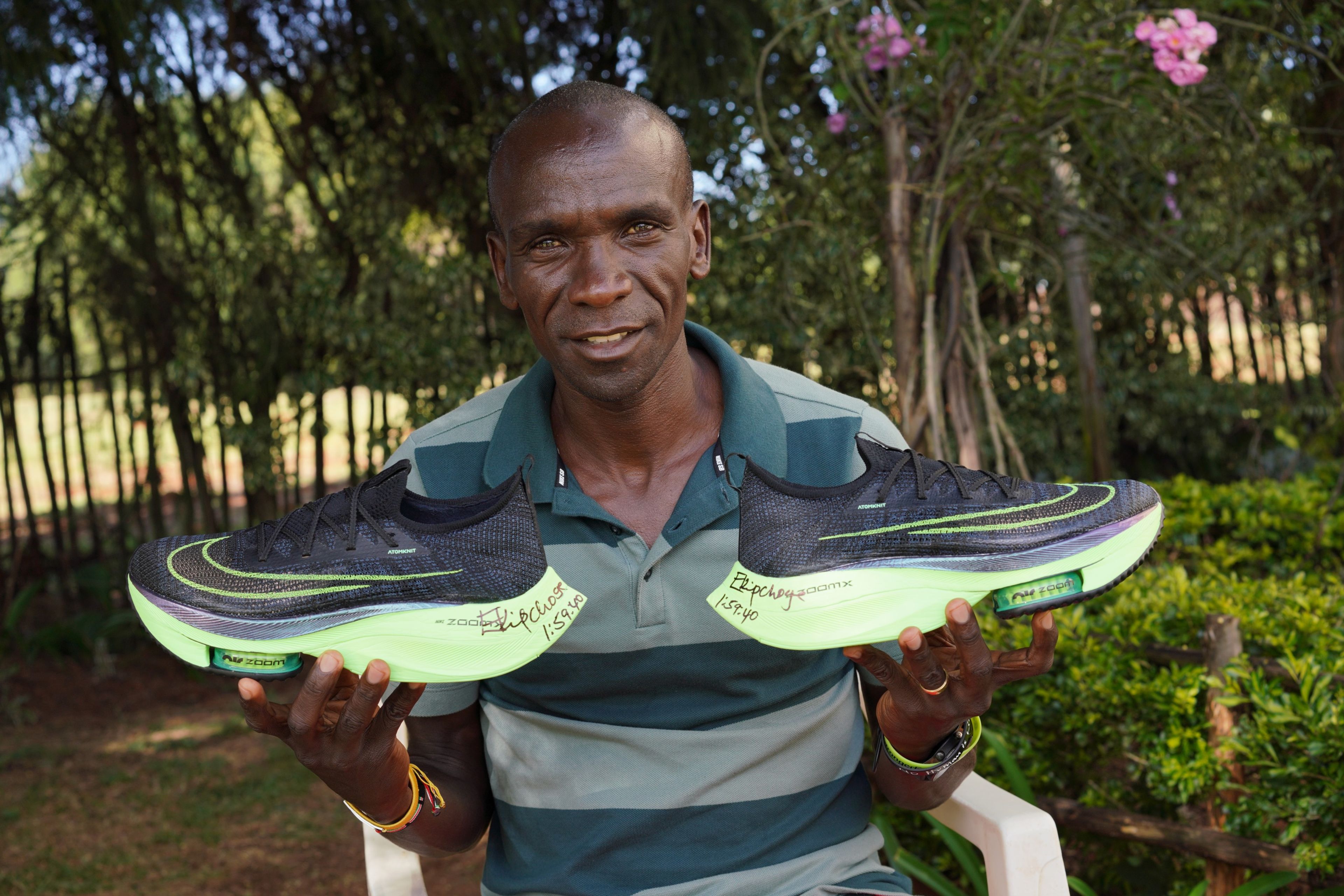 Eliud Kipchoge smiling and holding two signed Nike Air Zoom Alphafly shoes inscribed with his signature and 1:59:40, outdoors with green trees behind him.