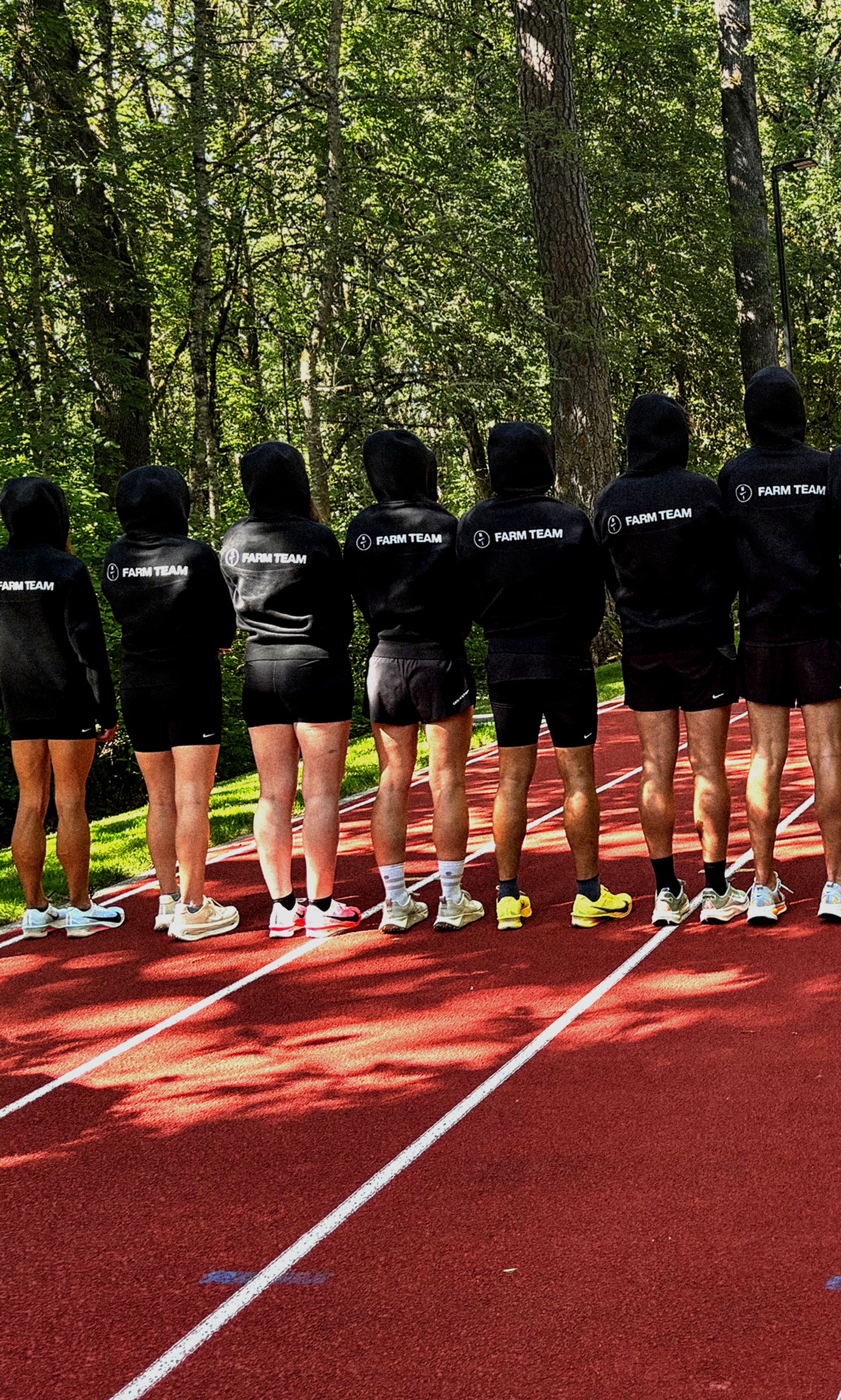 A line of athletes wearing black "FARM TEAM" hoodies and shorts stand on the Michael Johnson track facing away from the camera toward a forest.
