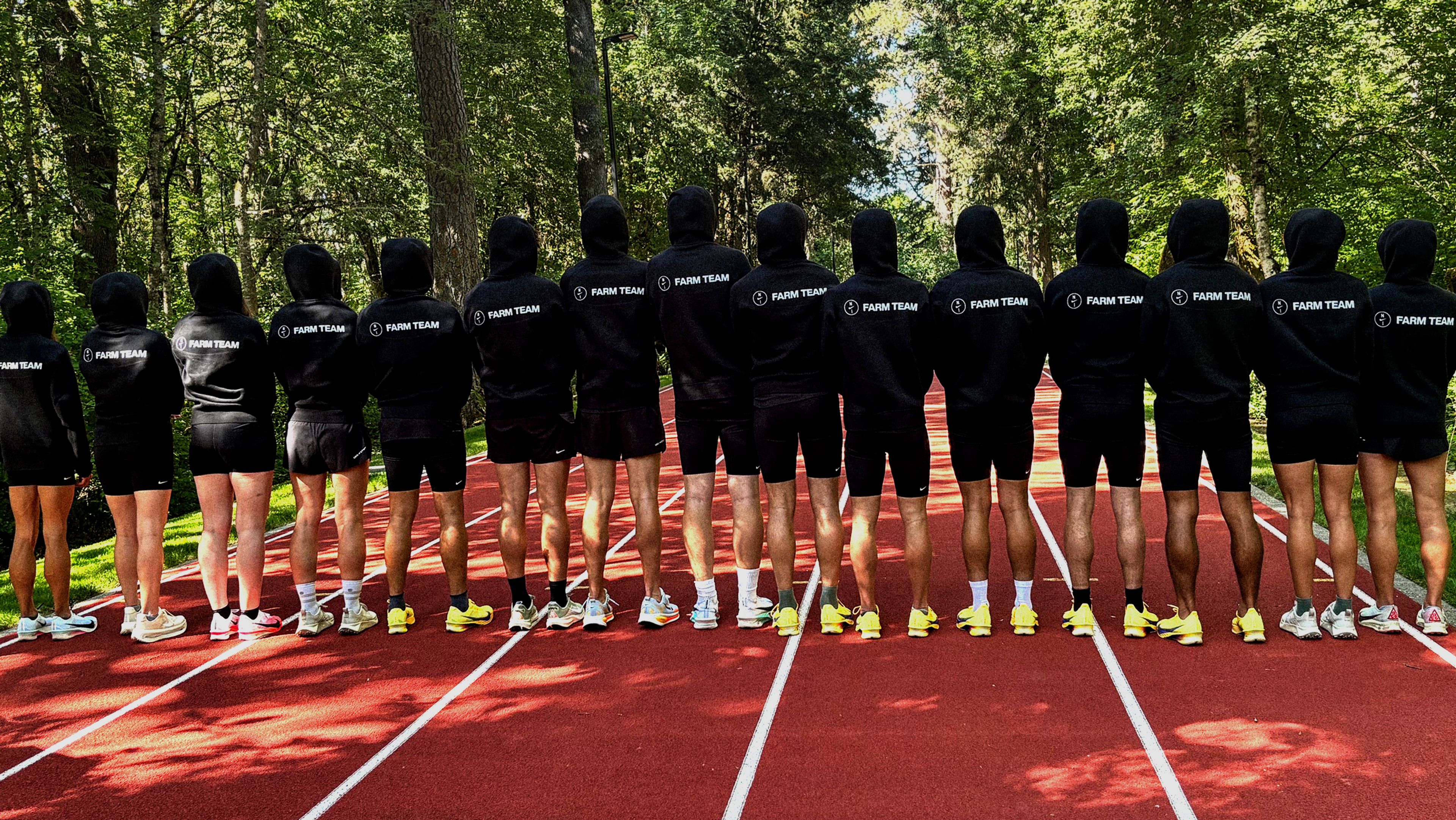 A line of athletes wearing black "FARM TEAM" hoodies and shorts stand on the Michael Johnson track facing away from the camera toward a forest.