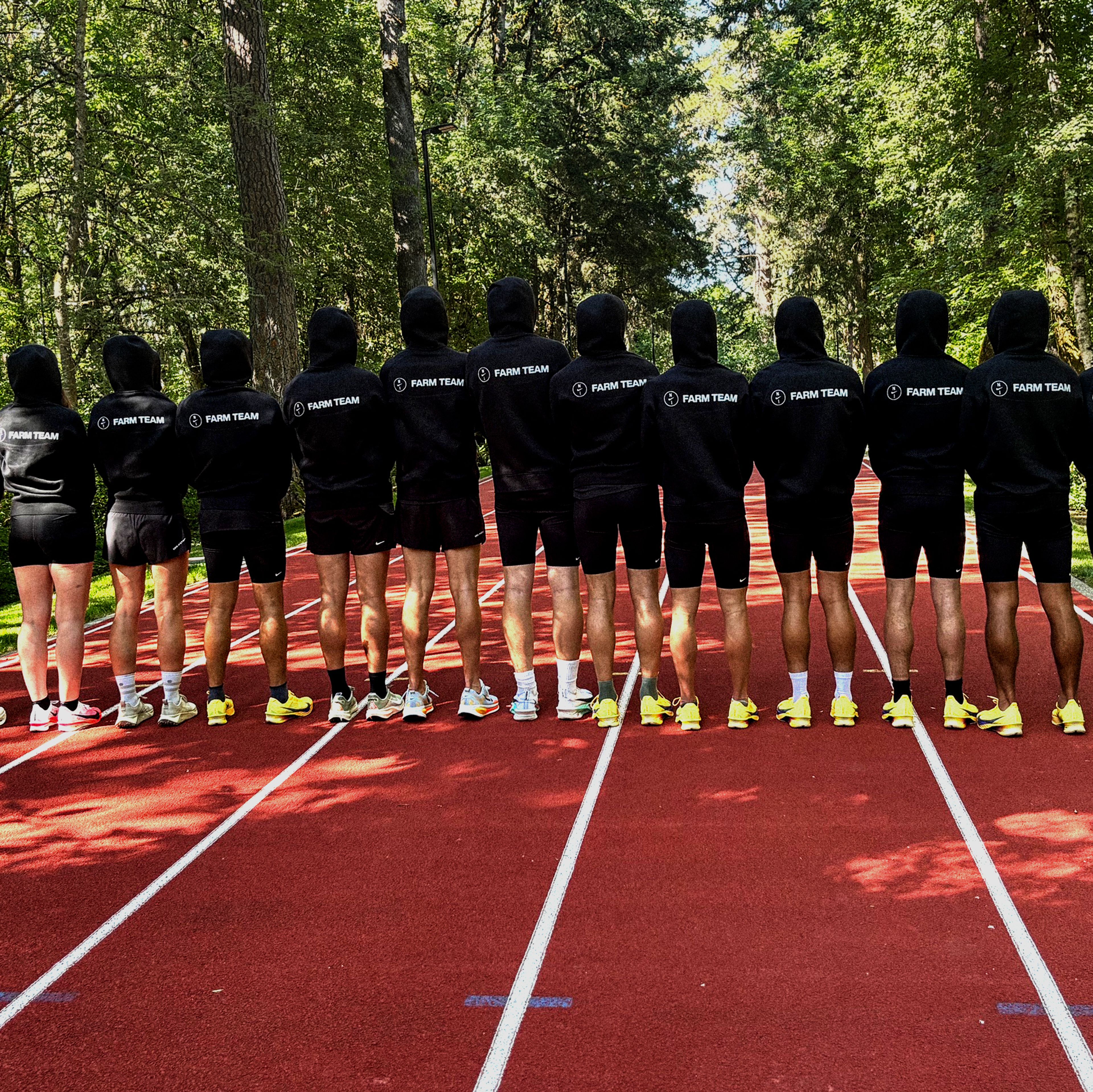 A line of athletes wearing black "FARM TEAM" hoodies and shorts stand on the Michael Johnson track facing away from the camera toward a forest.