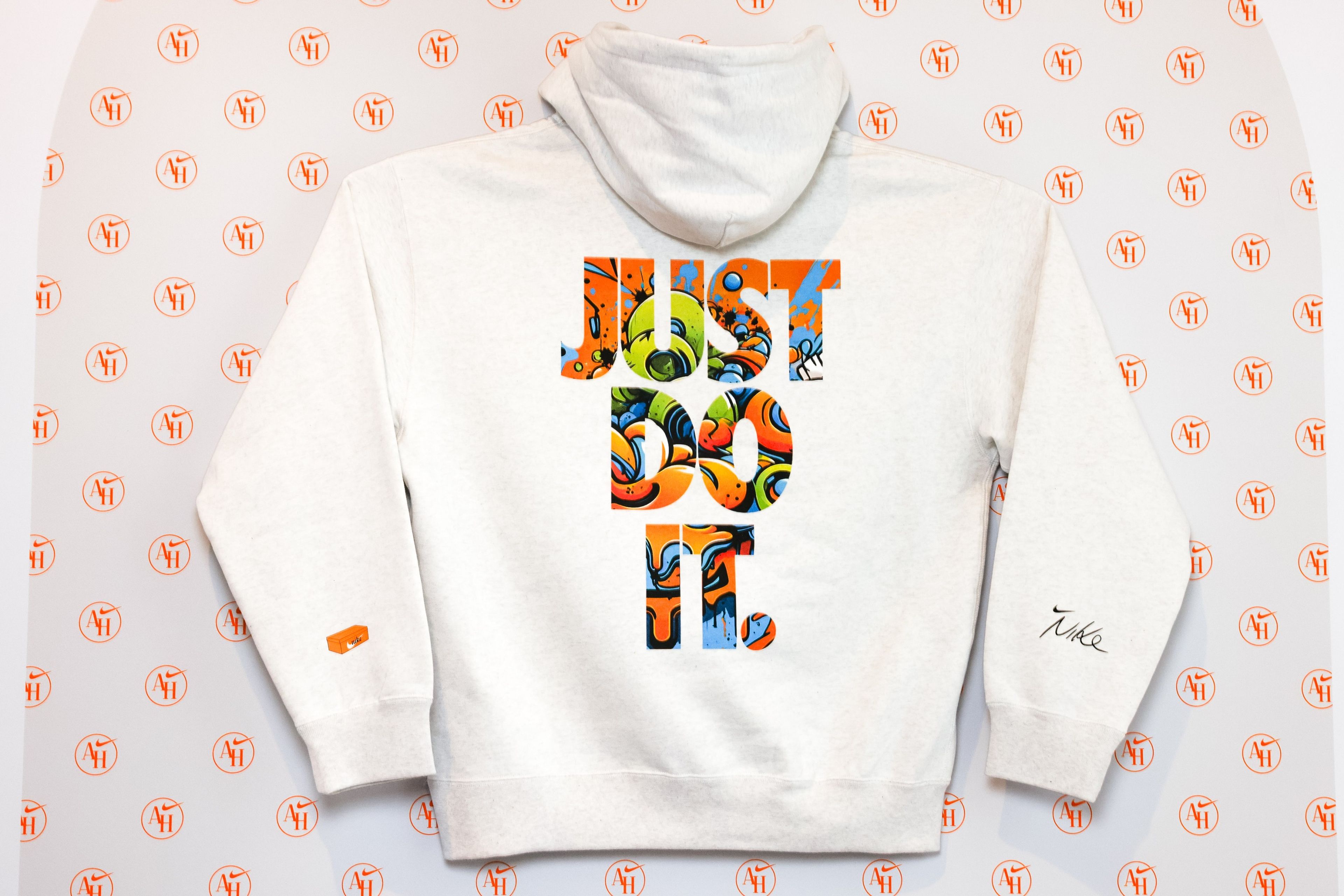 Back view of a white hoodie that has been customized in the AI space in the Nike Athlete House. A large, colorful JUST DO IT screenprint appears on the back of the sweatshirt, while the sleeves display a small orange footwear box and a scripted Nike logo. The hoodie sits on top of a repeating AH logo background.